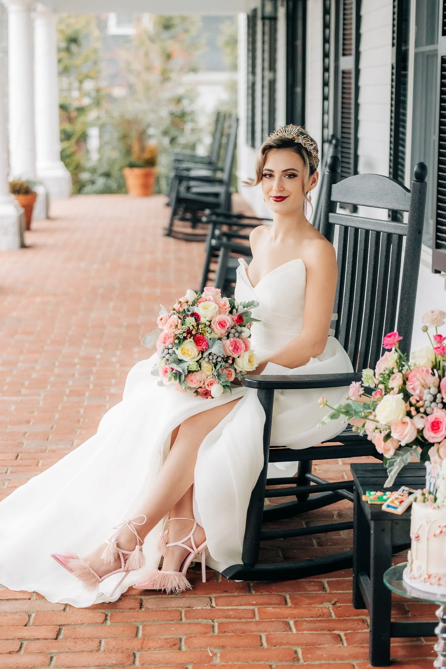 Bride in wedding dress holding bouquet, sitting on a black rocking chair on a brick porch.