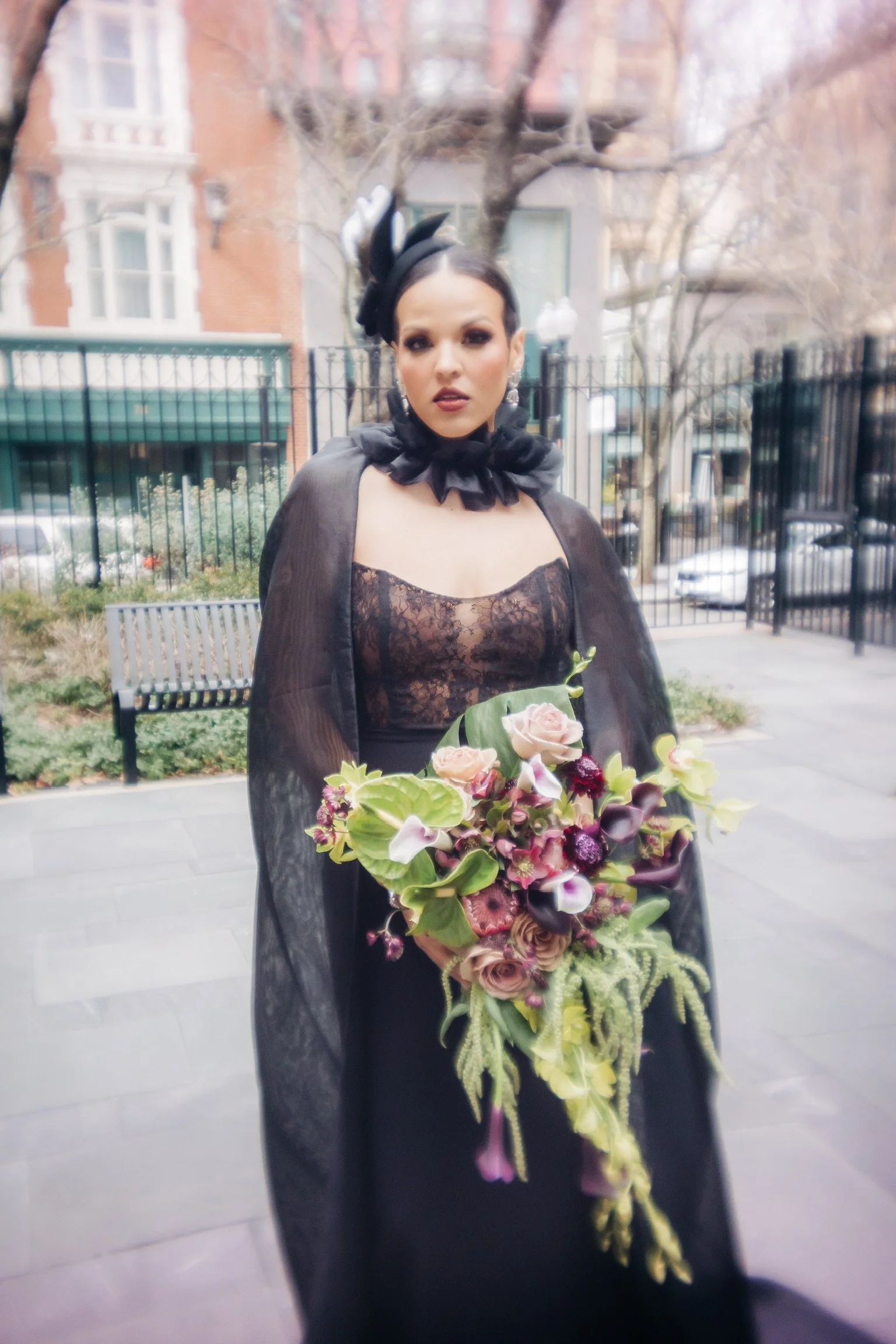 Woman in elegant black dress with floral bouquet, wearing a headpiece, standing outdoors.