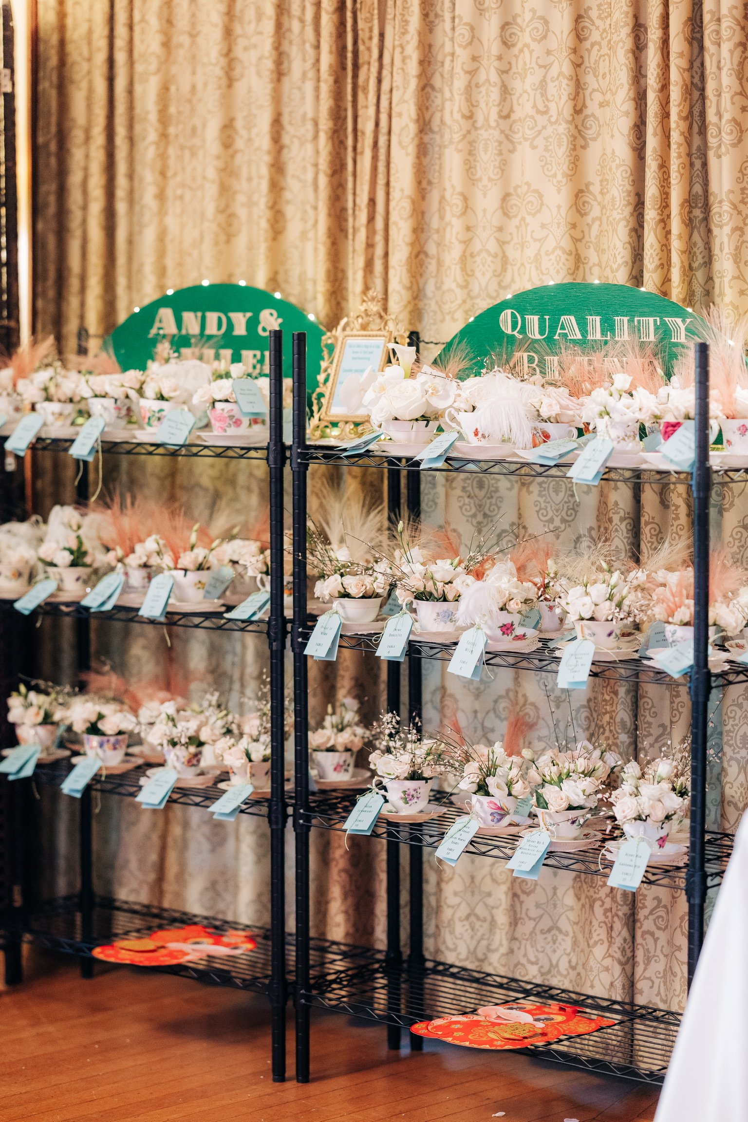 Shelves with floral arrangements in teacups, each tagged with a blue card, set against a patterned curtain background. Signs read 'Andy & Quality.'