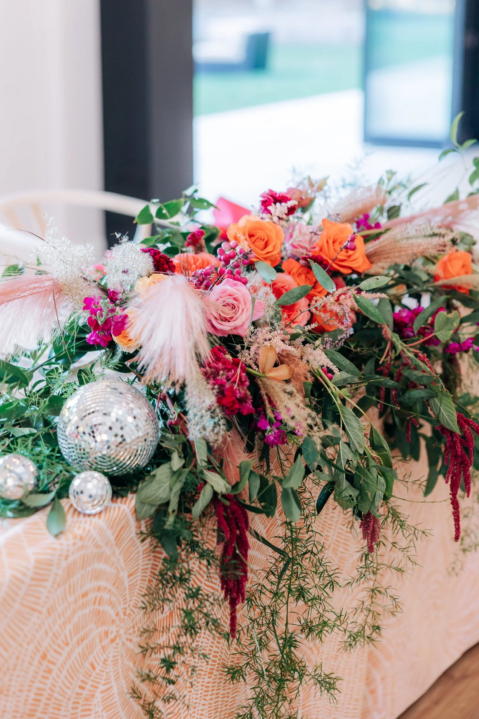 Floral arrangement with orange and pink roses, green foliage, pampas grass, and disco balls on a patterned tablecloth.