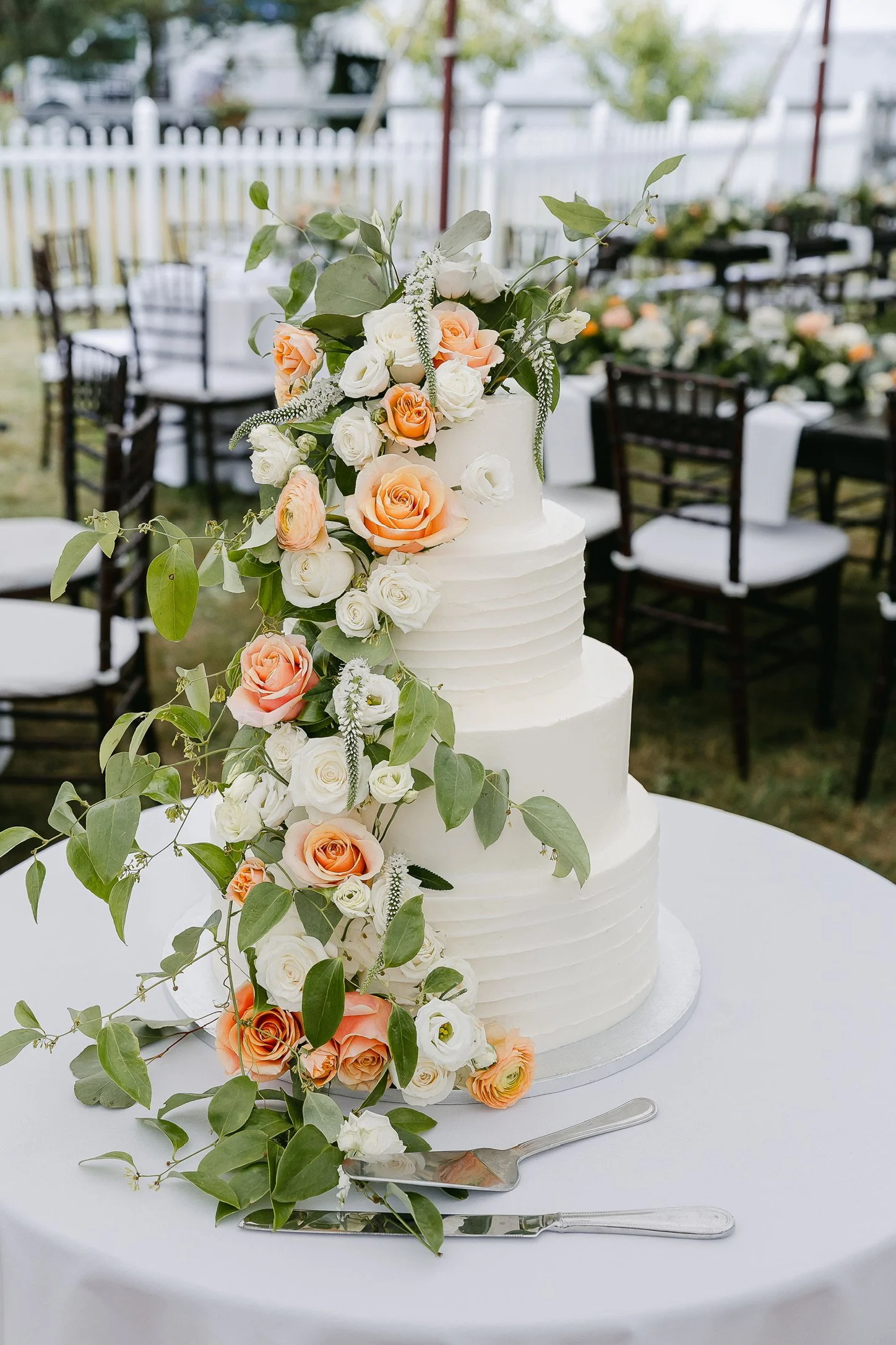 Three-tiered white wedding cake with peach and white roses, and green foliage, on a table with a white tablecloth, surrounded by chairs and a white fence in an outdoor setting.