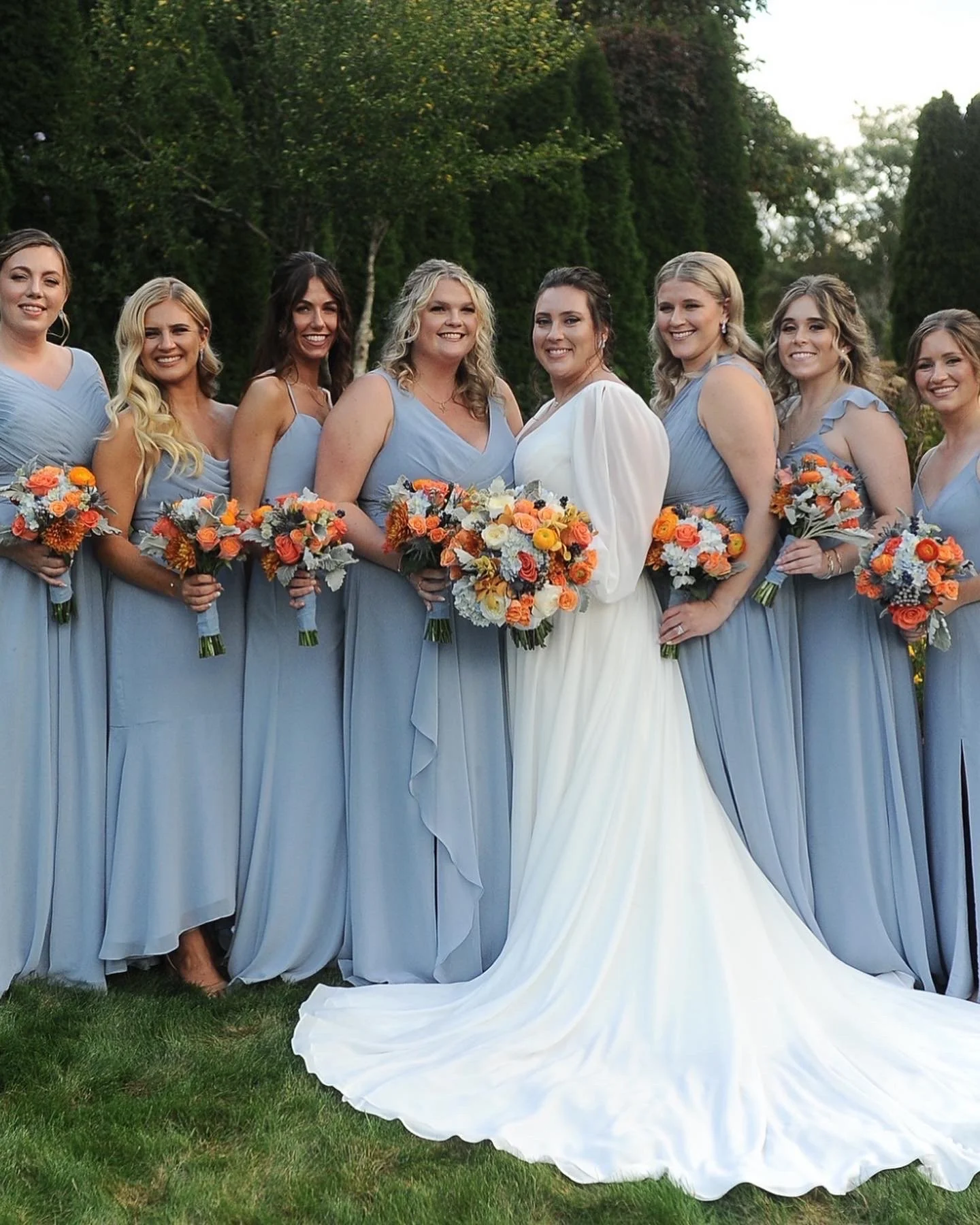 Bride and bridesmaids posing outdoors, bridesmaids in light blue dresses holding orange and white bouquets, and bride in a white gown.