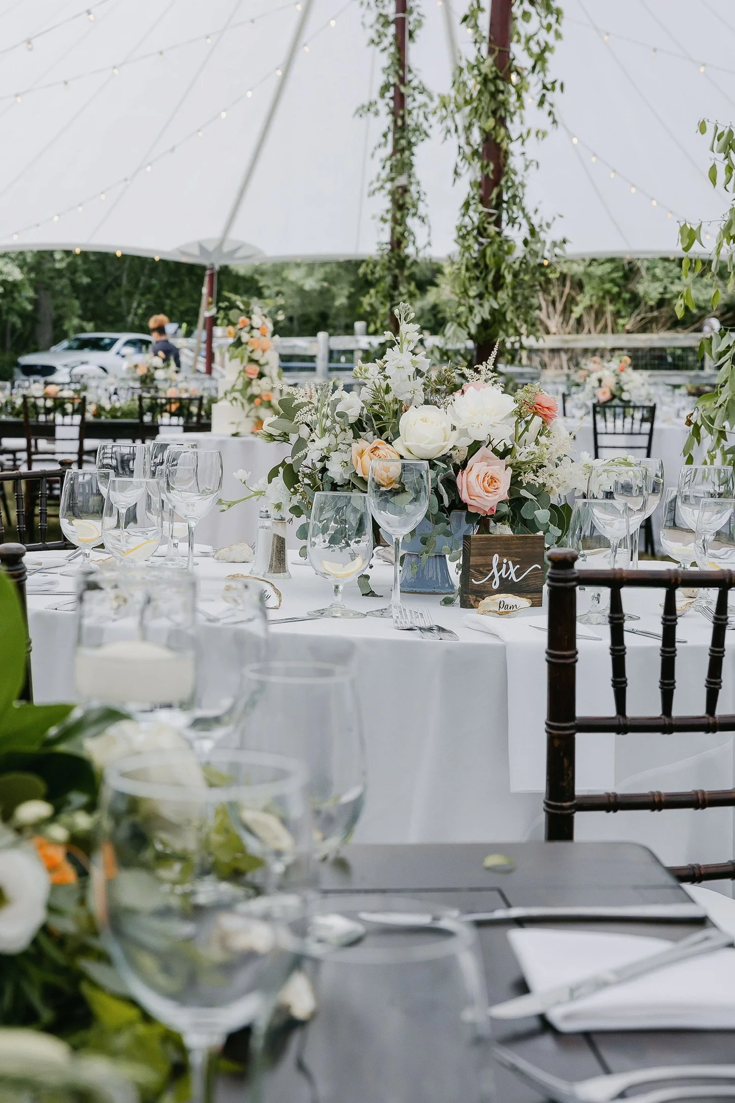 Outdoor wedding reception setup with round tables, white tablecloths, floral centerpieces, and string lights under a tent.