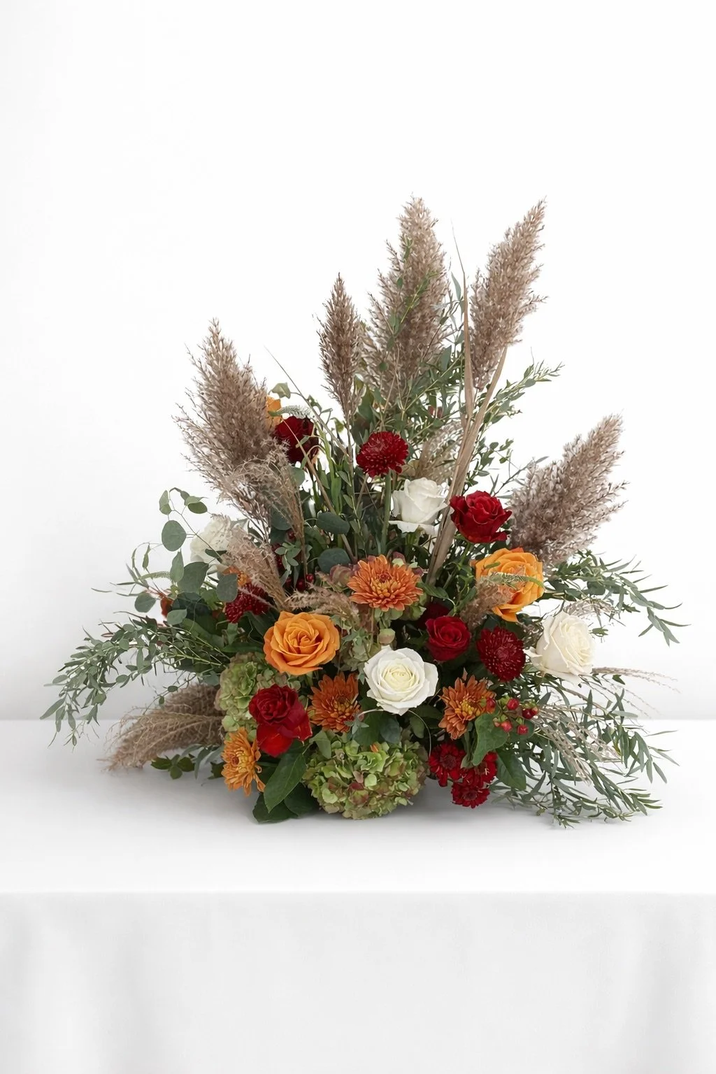 A floral arrangement with orange roses, red roses, white roses, orange dahlias, red carnations, green hydrangeas, and beige pampas grass, displayed against a plain white background.