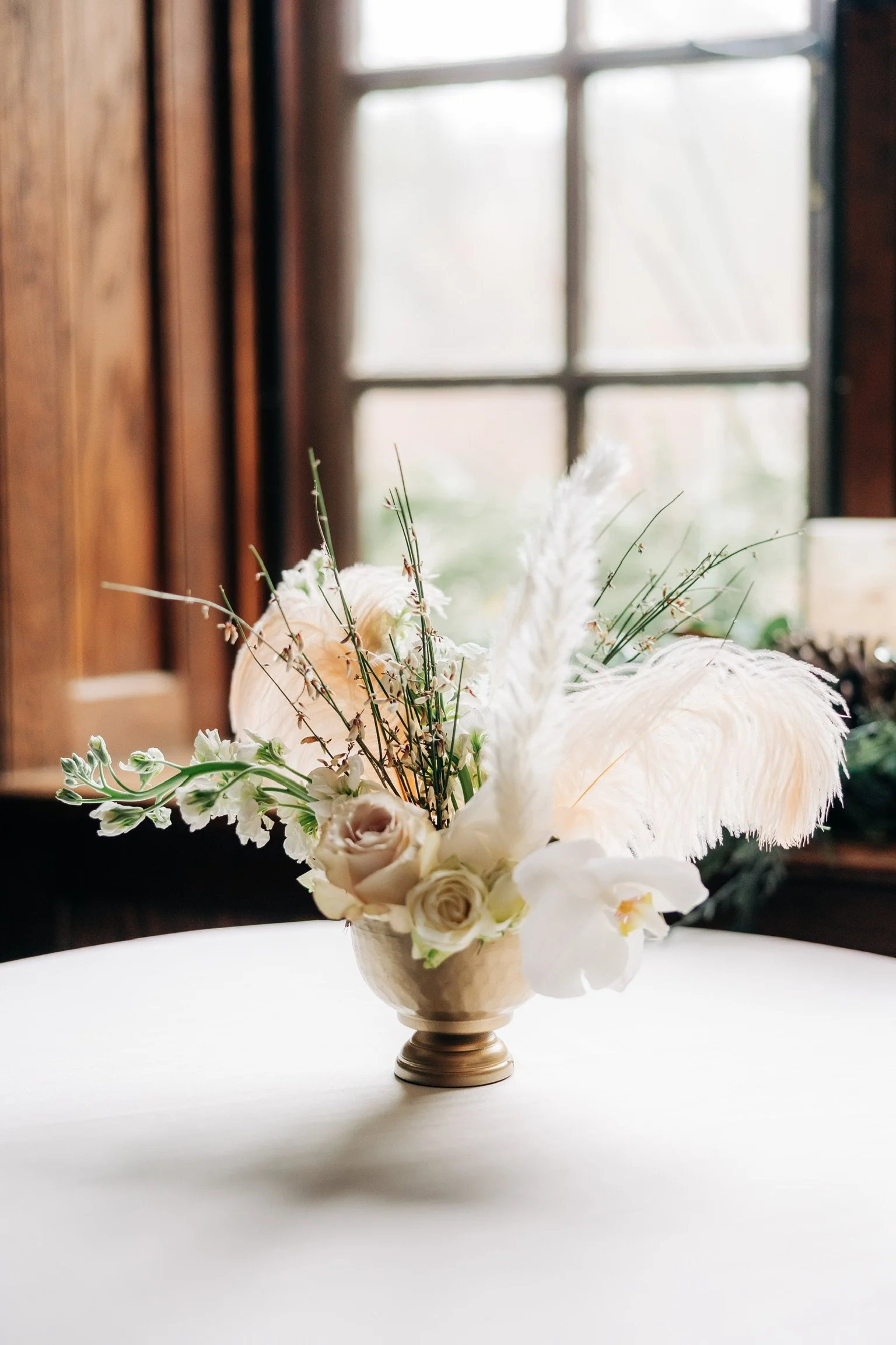 Elegant floral arrangement with roses, feathers, and branches in a gold vase on a table.