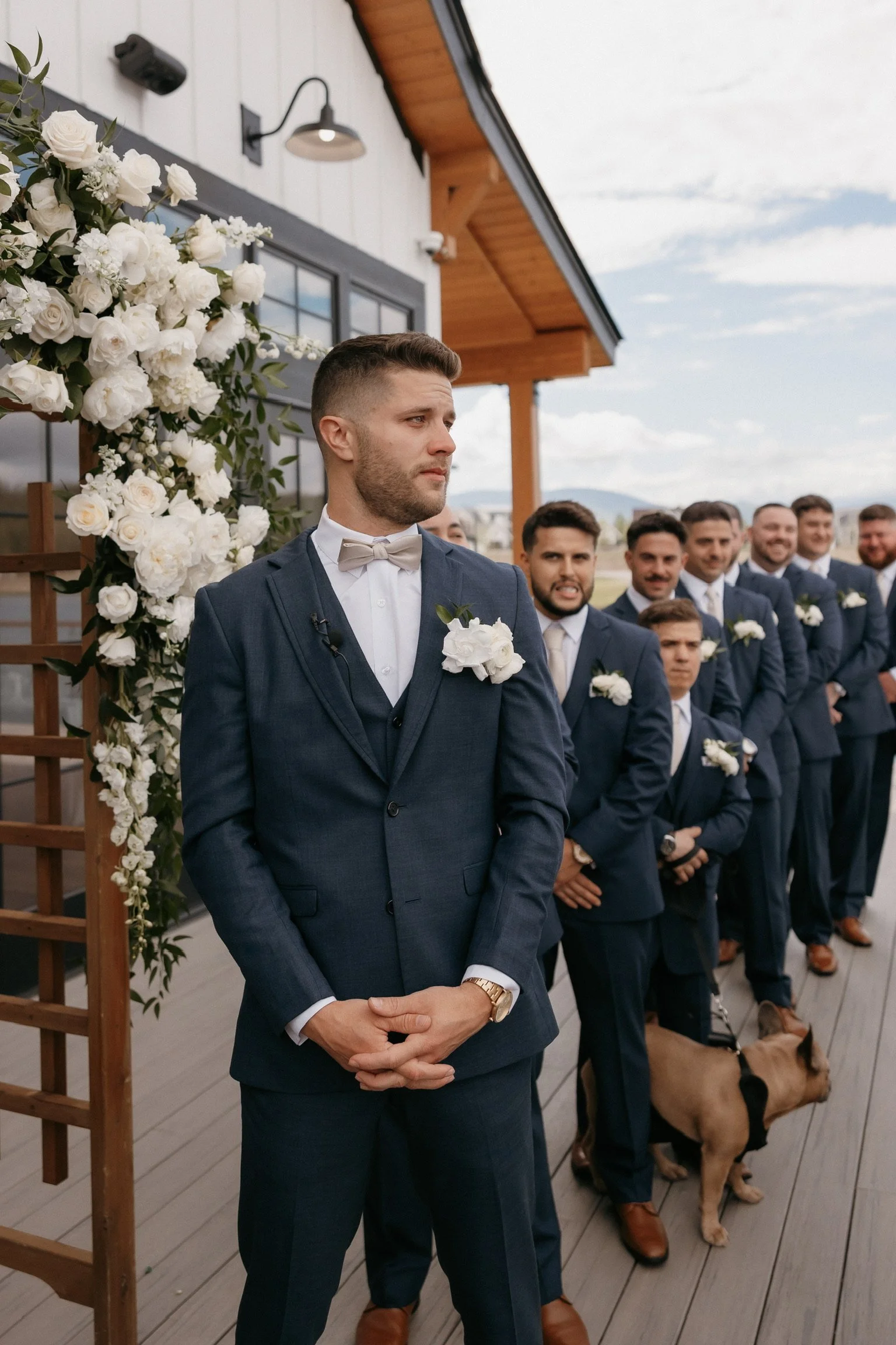 Groomsmen lined up outside a building, wearing navy suits with white boutonnieres. A floral arch with white flowers is on the left. A dog is sitting near the front groomsman.