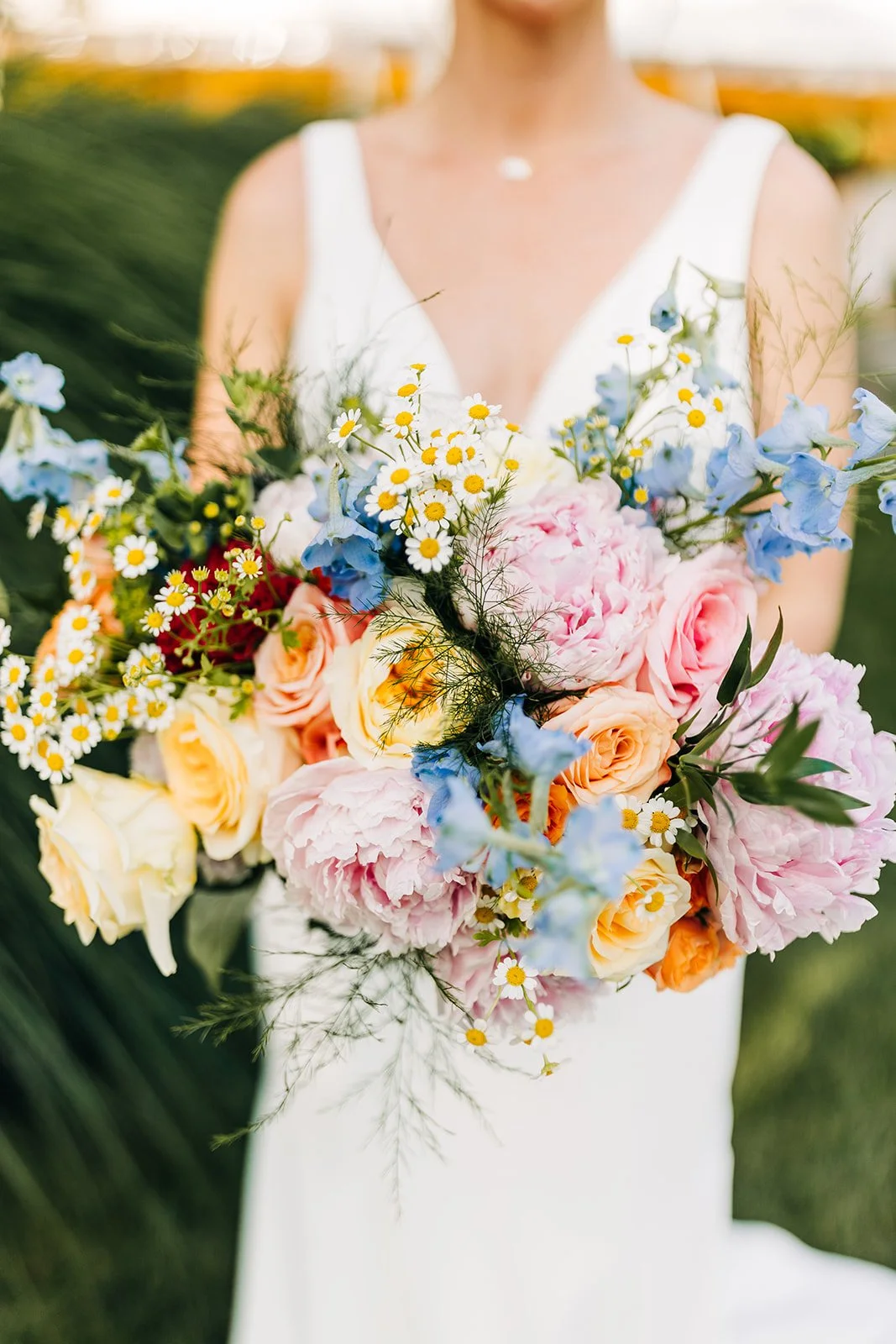 Bride holding colorful bouquet with pink peonies, yellow roses, blue delphiniums, and daisies.