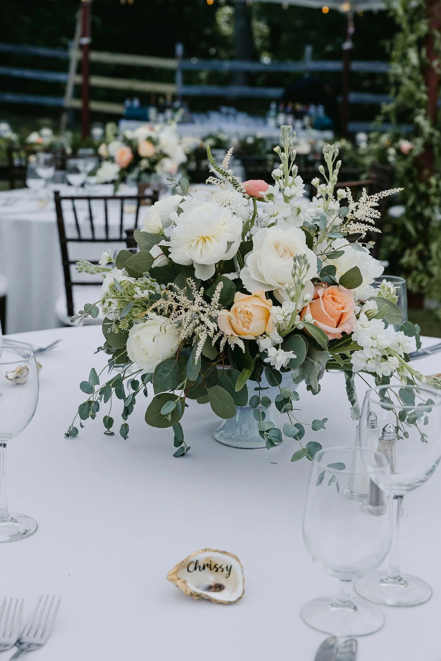 Elegant table setting with a floral centerpiece and glassware, featuring white and peach roses, green foliage, and a decorative name card shell.