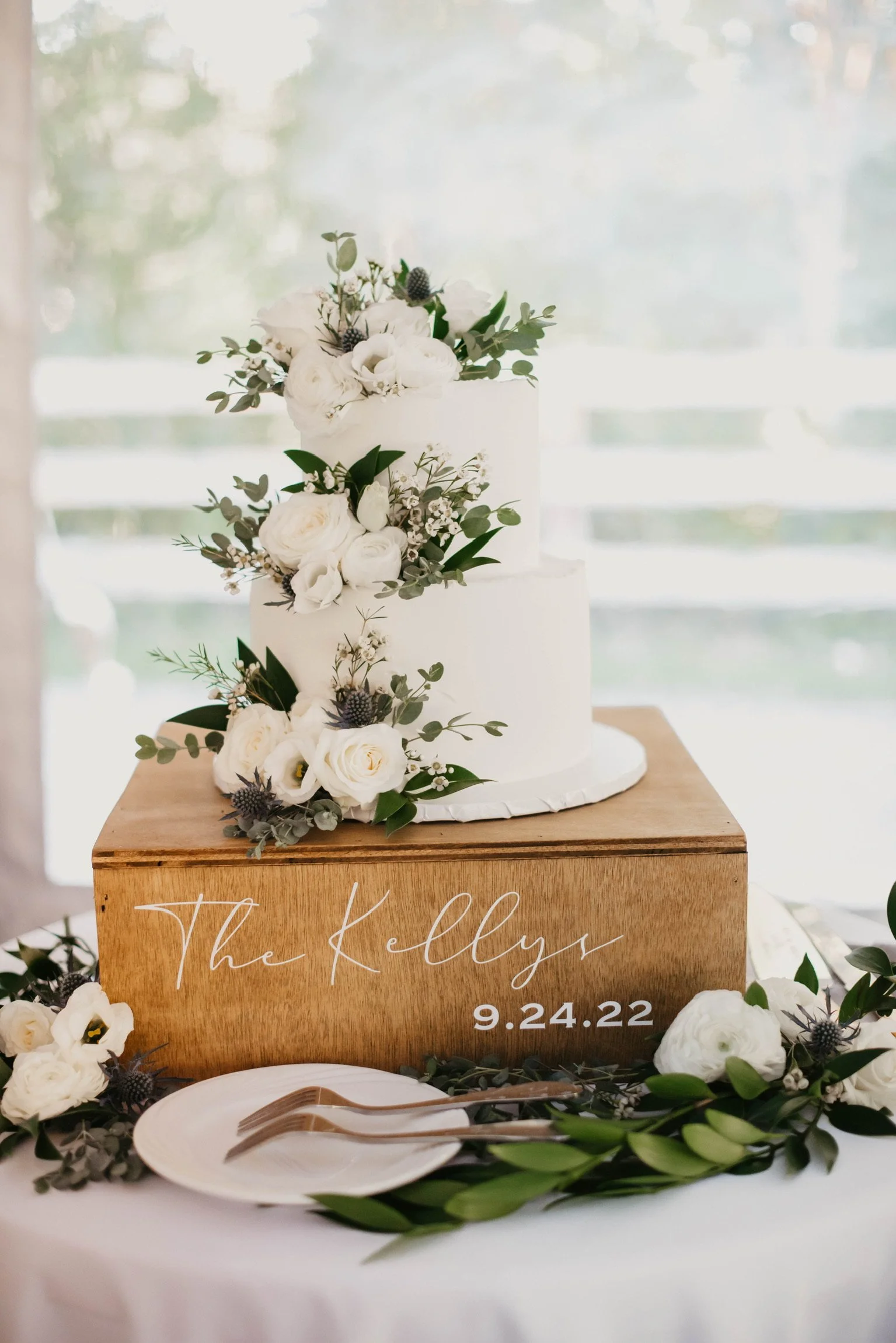 Two-tier white wedding cake with floral decorations on a wooden box labeled "The Kellys 9.24.22" with plates and forks nearby.