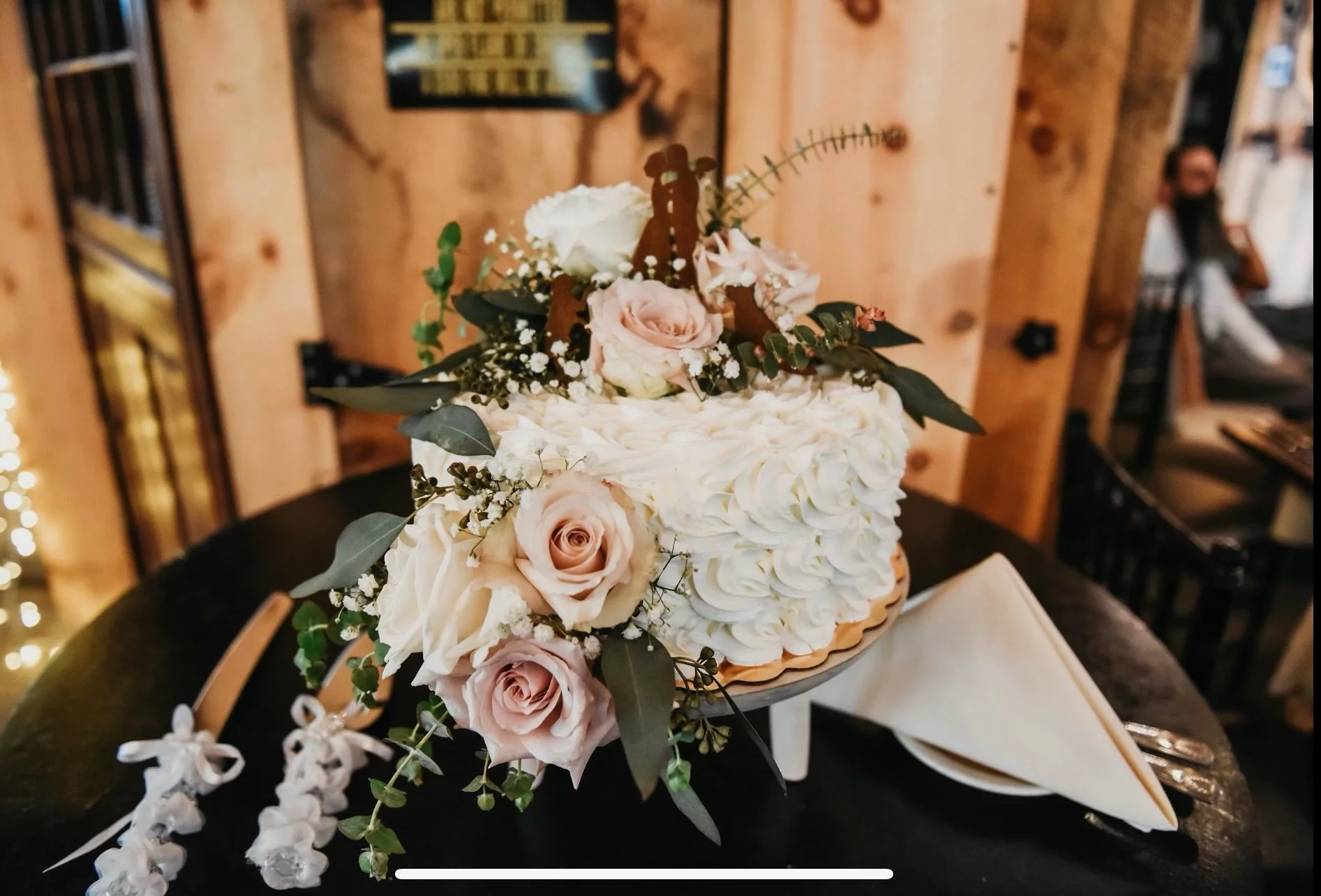 A wedding cake with white rose frosting, topped with pink and white roses, eucalyptus leaves, and baby's breath, on a wooden stand. The cake is accompanied by a cake knife and server with decorative bows.