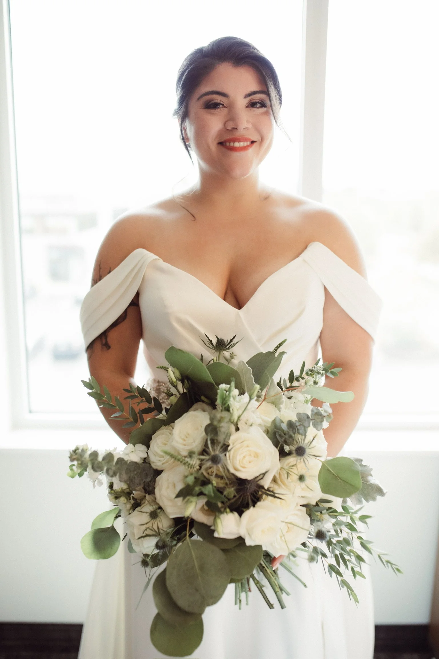 Bride in off-shoulder white dress holding a bouquet of white roses and greenery, standing in front of a window.