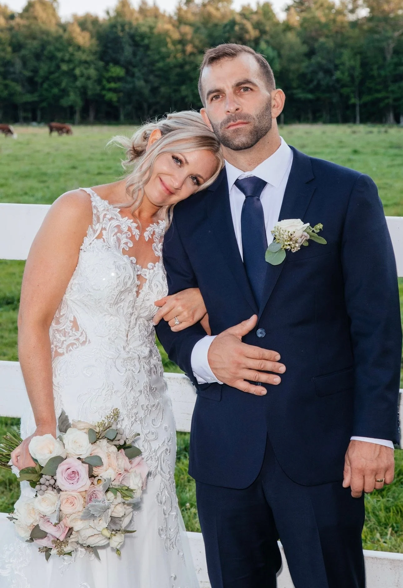A couple in wedding attire posing by a white fence with a field and trees in the background. The bride is wearing a white lace gown and holding a bouquet of roses, while the groom is in a dark suit with a flower boutonniere.