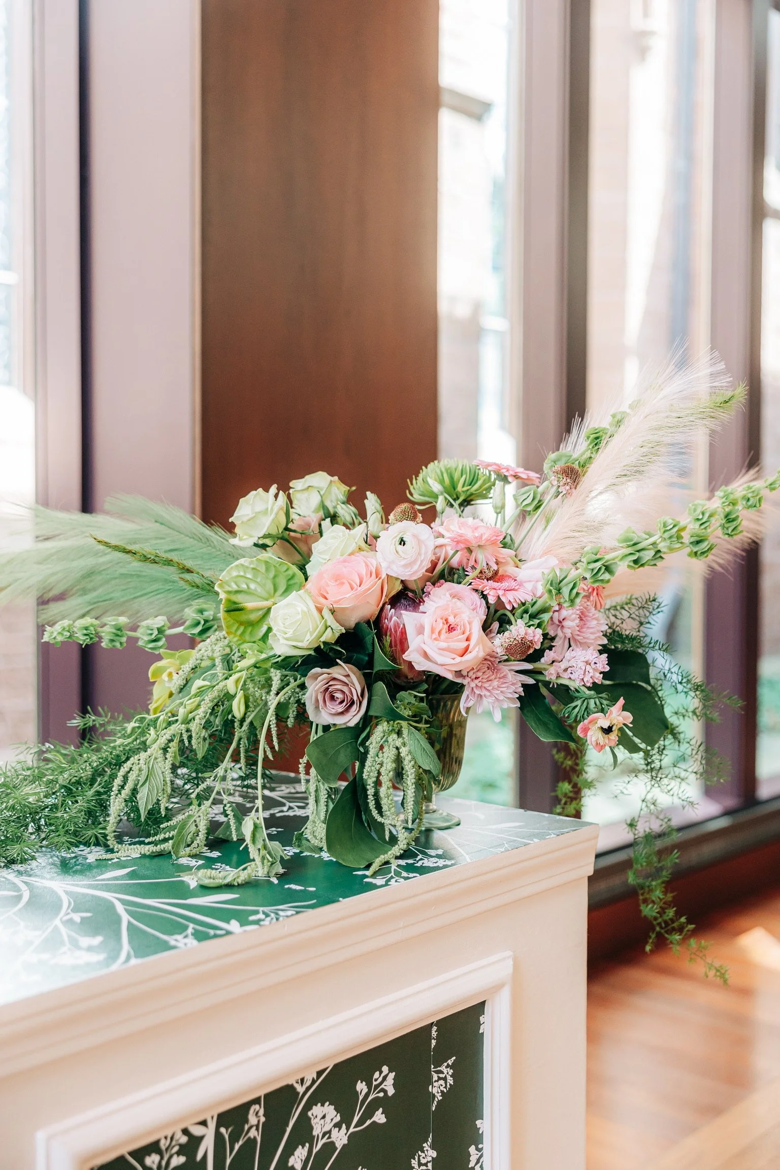 Elegant floral arrangement with pink roses, green leaves, and assorted blooms on a decorated surface near large windows.