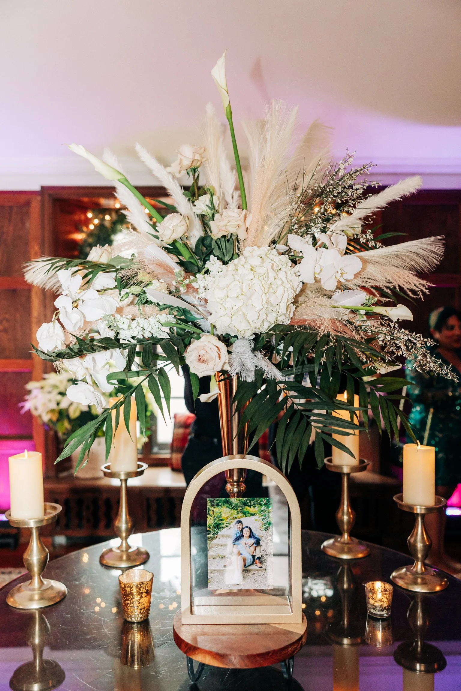 Elegant floral arrangement with white and green flowers, pampas grass, and foliage, placed on a table with candles and a framed photo.