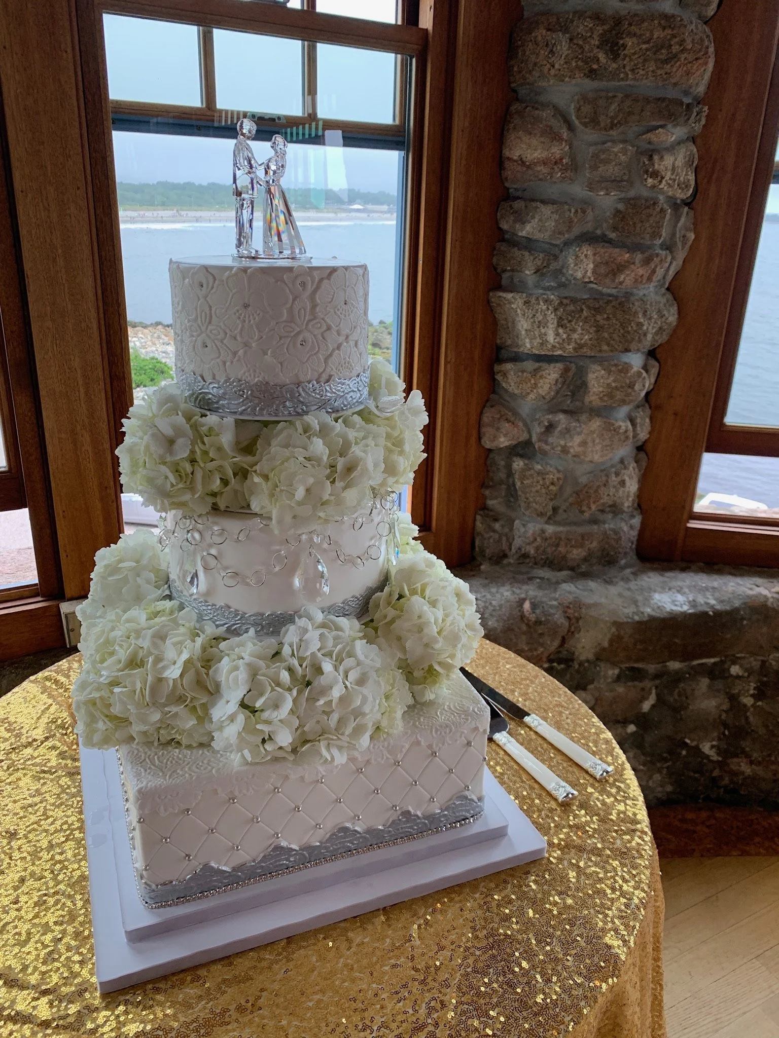 A three-tiered white wedding cake decorated with intricate patterns, white flowers, and silver accents, topped with a crystal bride and groom figurine, placed on a gold-sequined tablecloth by a window with a waterfront view.
