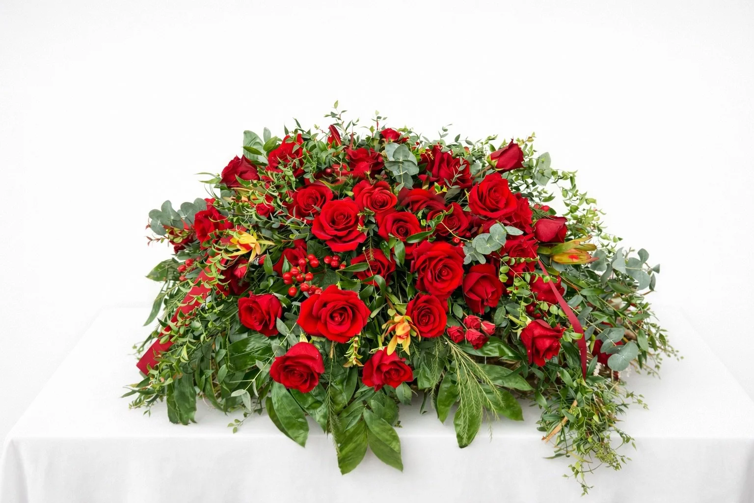 A large floral arrangement of red roses, green leaves, and some small red berries on a white table against a white background.