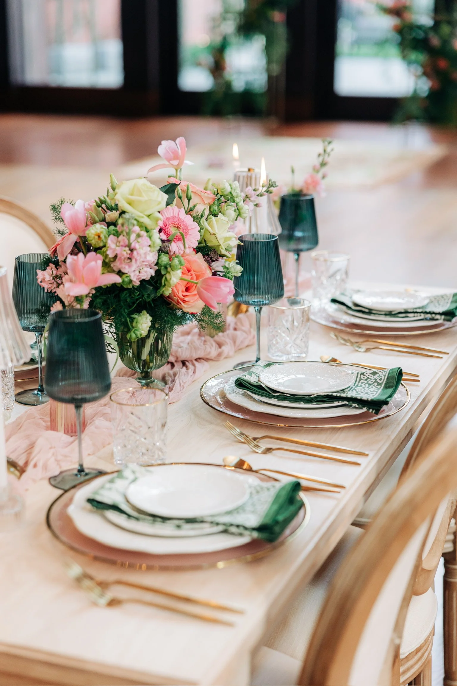 Elegant dining table set with floral centerpiece, green glassware, white and green table settings, gold cutlery, and textured napkins.