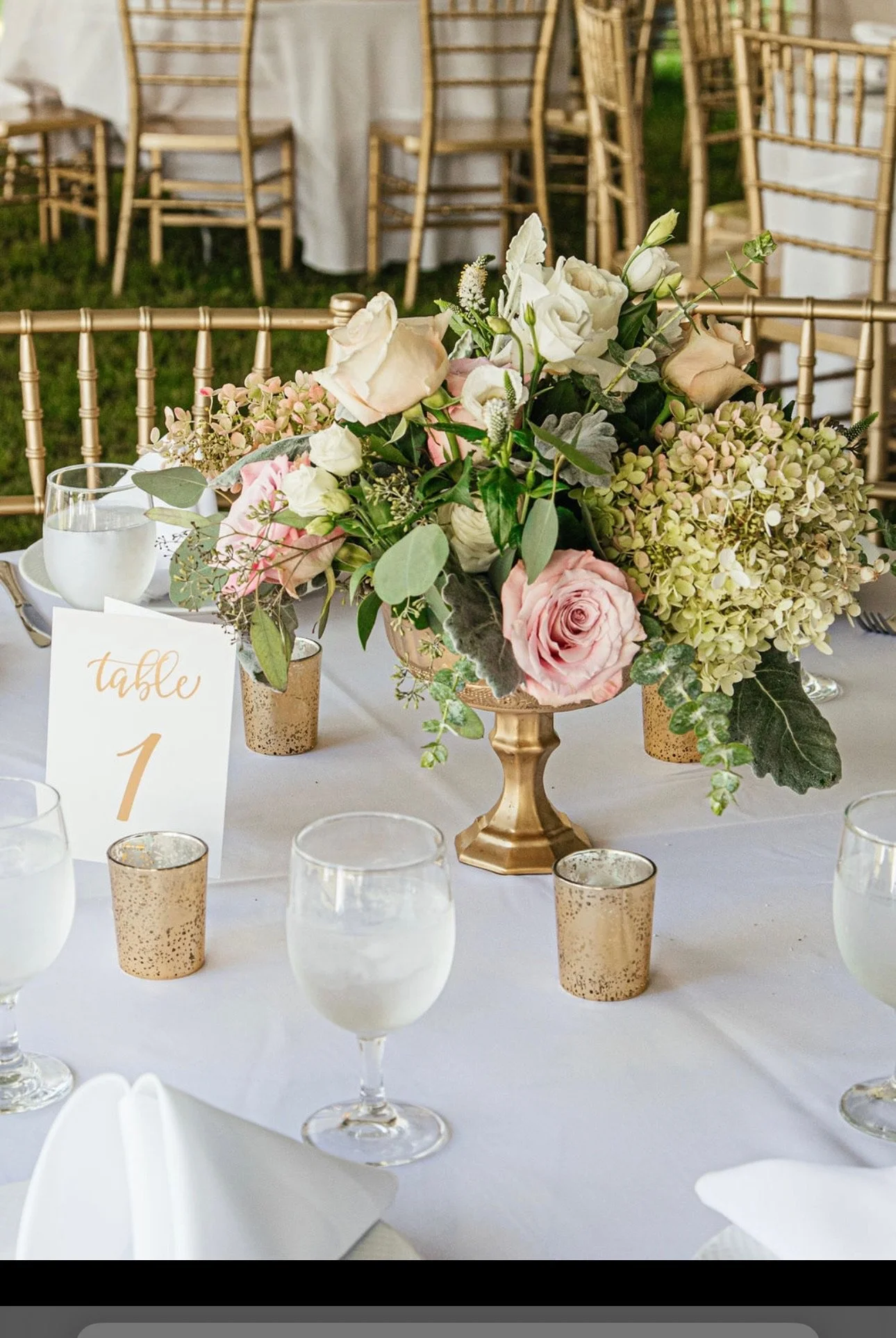 Table setting with centerpiece of pink roses, white flowers, and greenery in a gold vase, surrounded by gold votive candle holders, glasses of water, and a "Table 1" card.