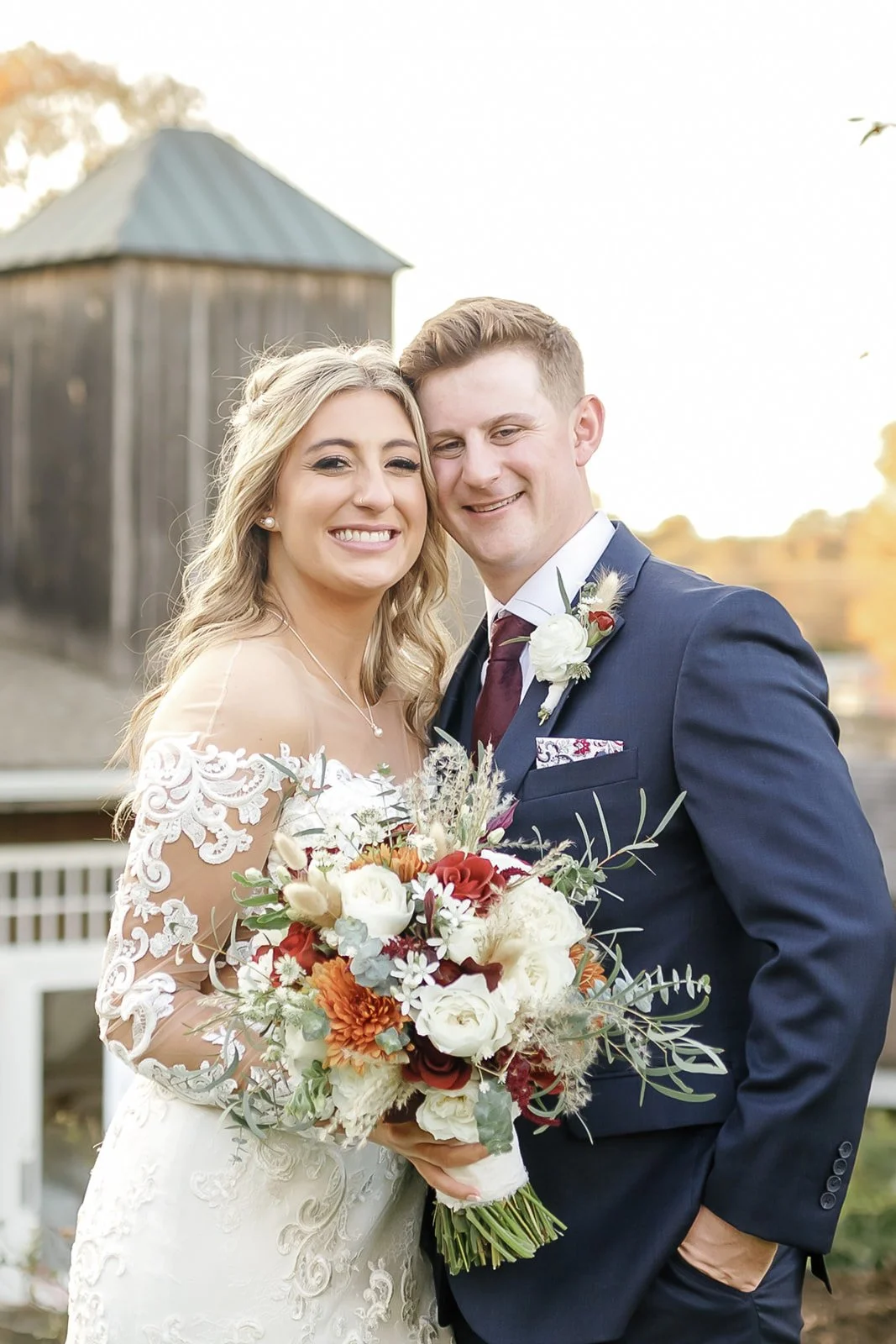A bride and groom smiling, posing for a wedding photo outdoors, with a rustic barn in the background. The bride holds a bouquet of flowers.