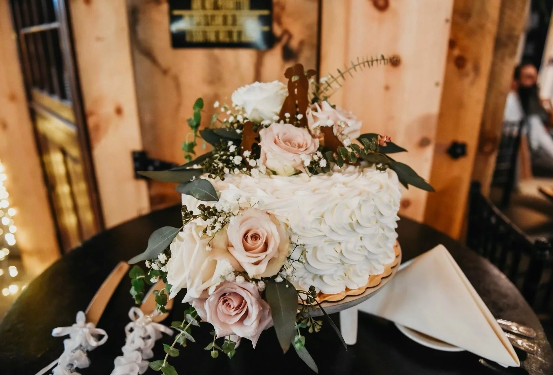 White frosted cake with pink roses and greenery on top, placed on a round table with utensils and napkins.