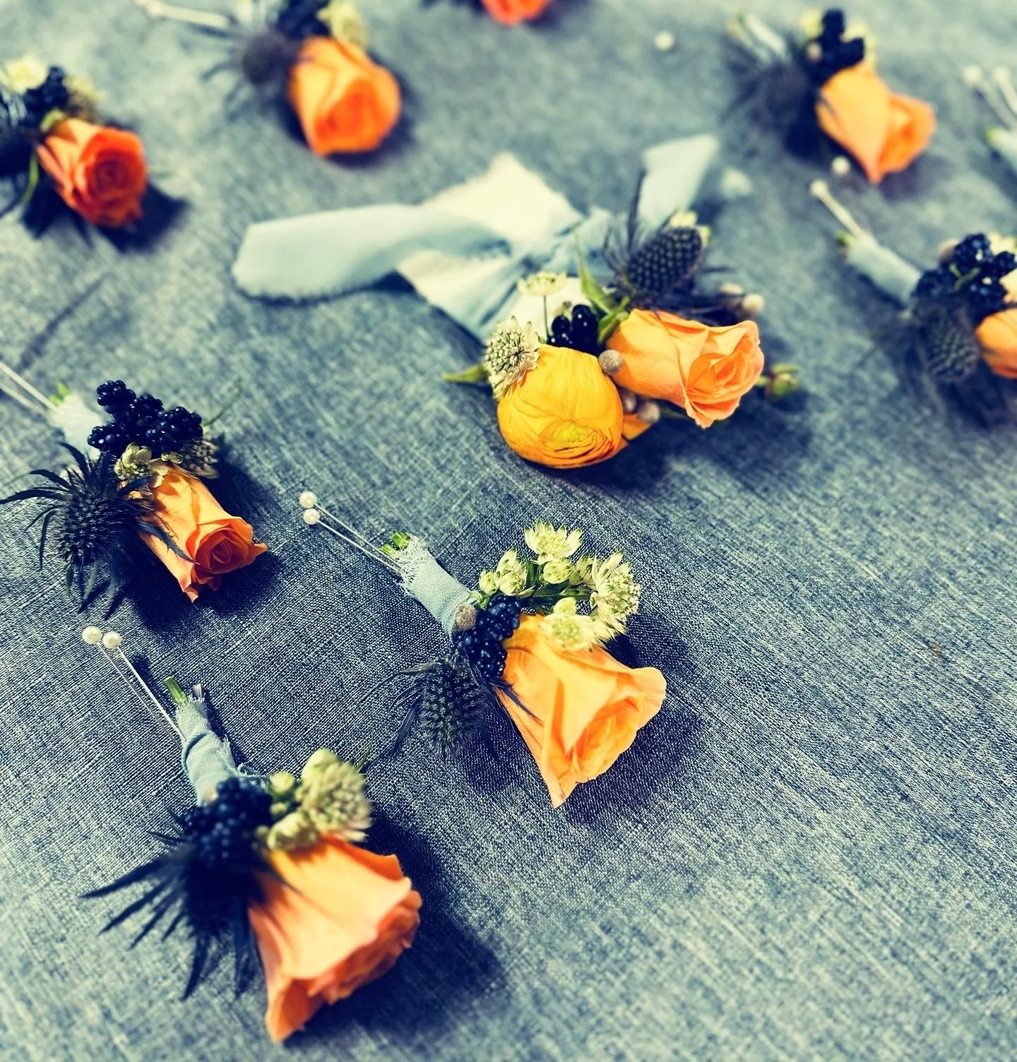 Orange rose boutonnieres and small bouquets arranged on a gray fabric, with decorative berries and greenery.