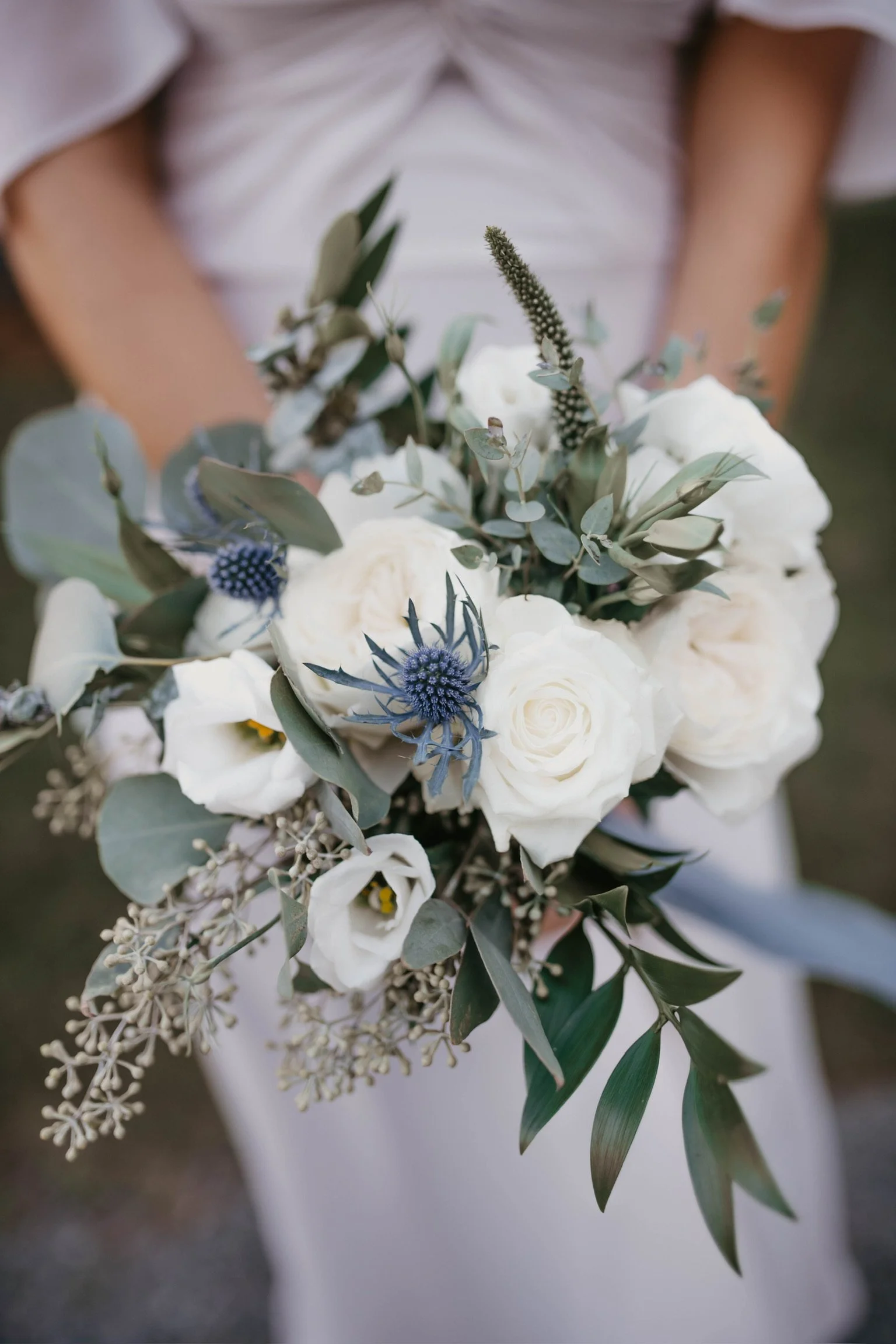 Close-up of a wedding bouquet with white roses, blue thistles, eucalyptus leaves, and greenery.