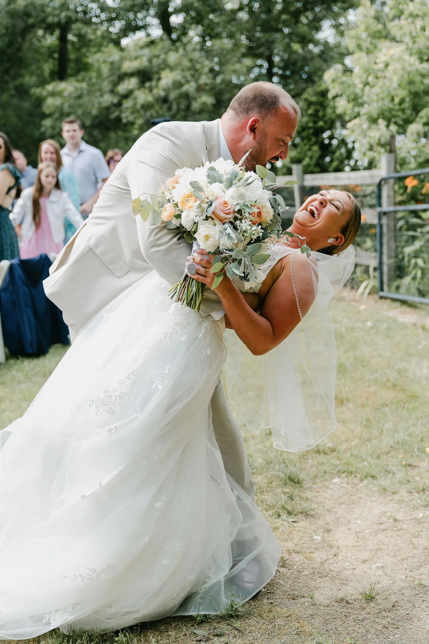 Wedding couple laughing and dancing outdoors, bride holding a bouquet.