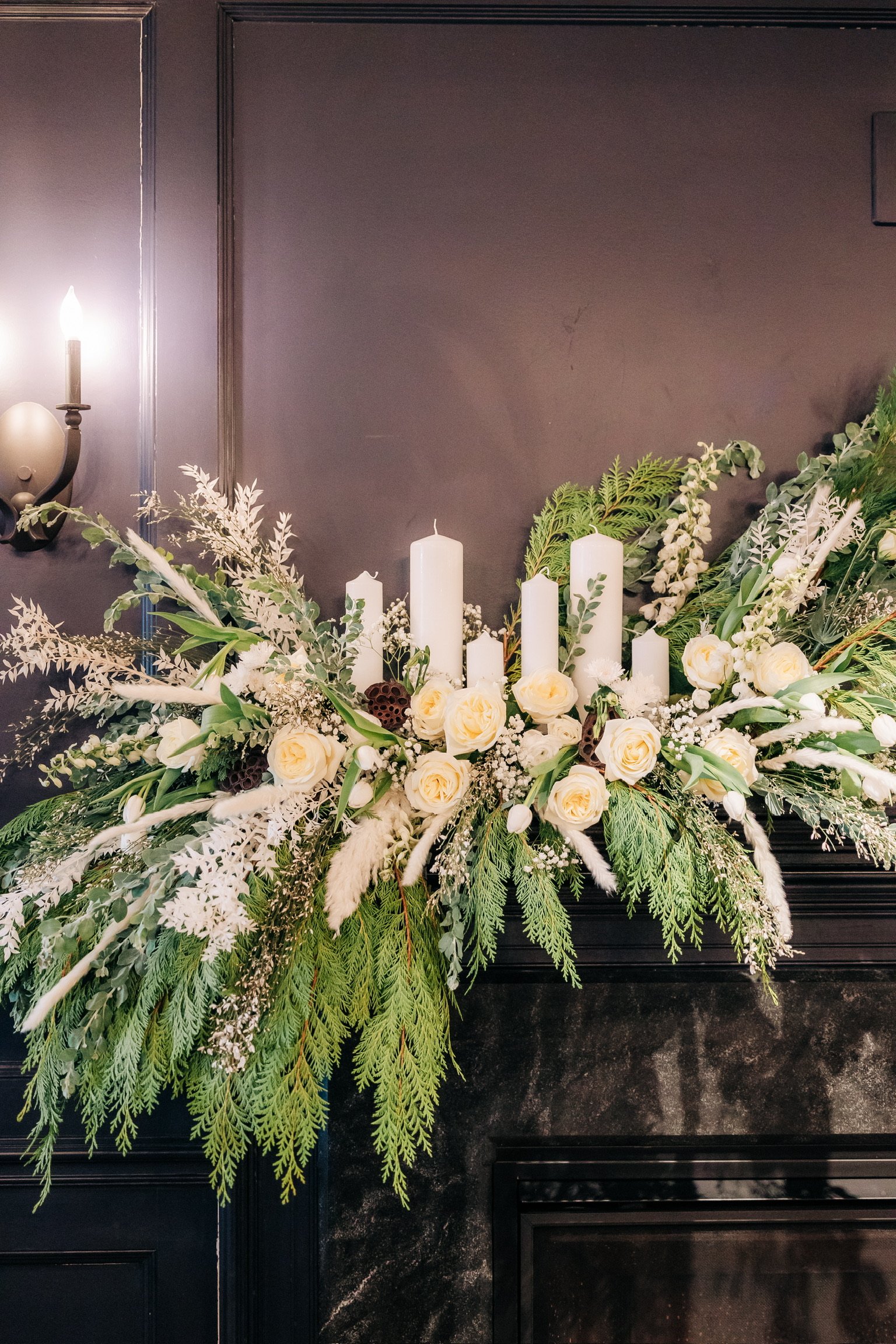 Elegant floral arrangement with white roses, greenery, and tall white candles on a dark mantel.