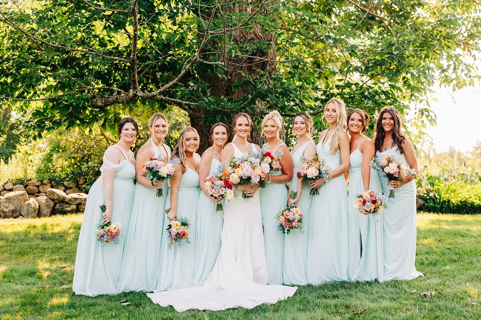 Bridal party with bride and bridesmaids wearing light blue dresses holding bouquets in a garden setting.