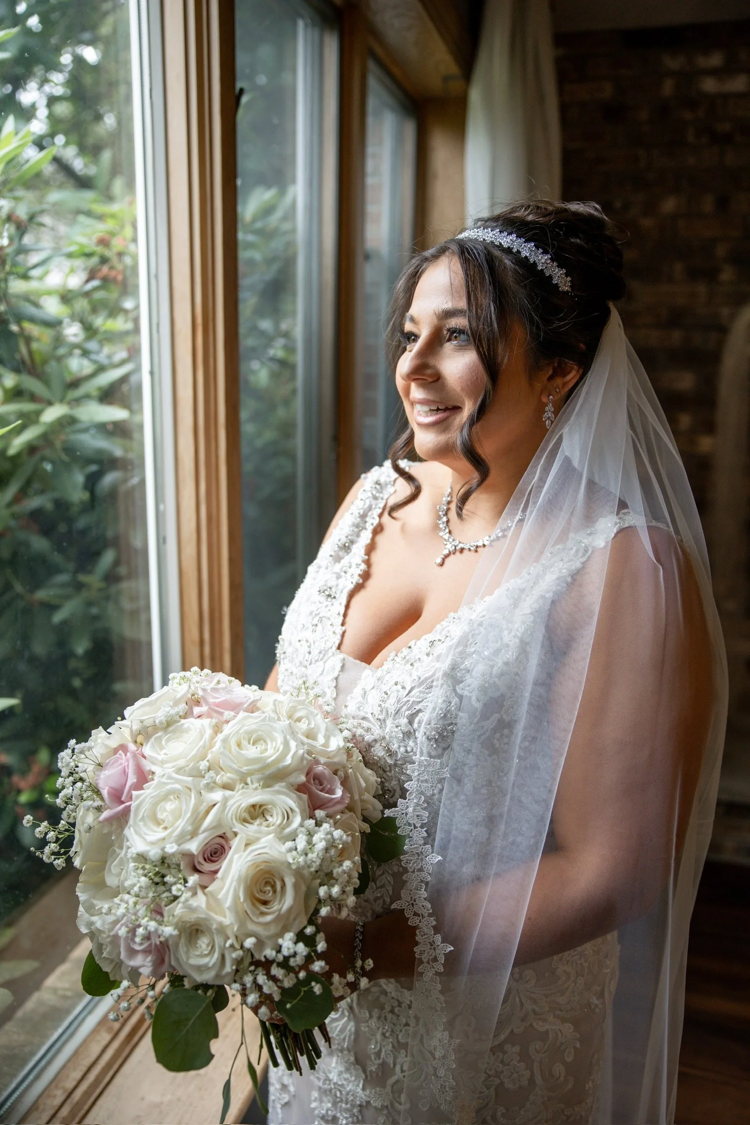Bride in lace wedding dress holding a bouquet of white and pink roses, looking out of a window, wearing a veil and jeweled headpiece.