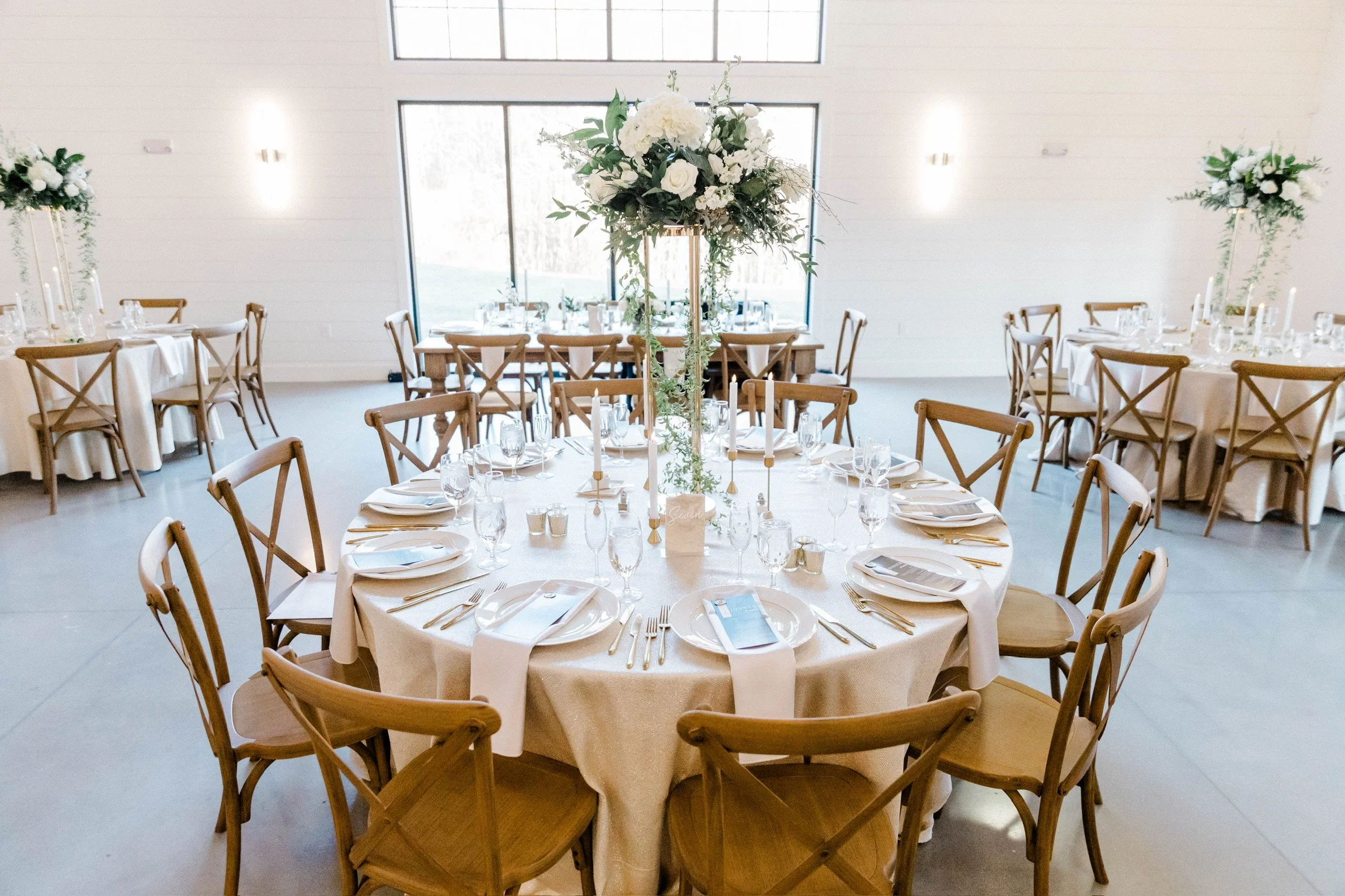 Elegant banquet hall with round tables covered in white tablecloths, set with white plates, silverware, and wine glasses. Tall floral centerpieces with white flowers and greenery decorate the tables. Wooden cross-back chairs surround the tables. Larg