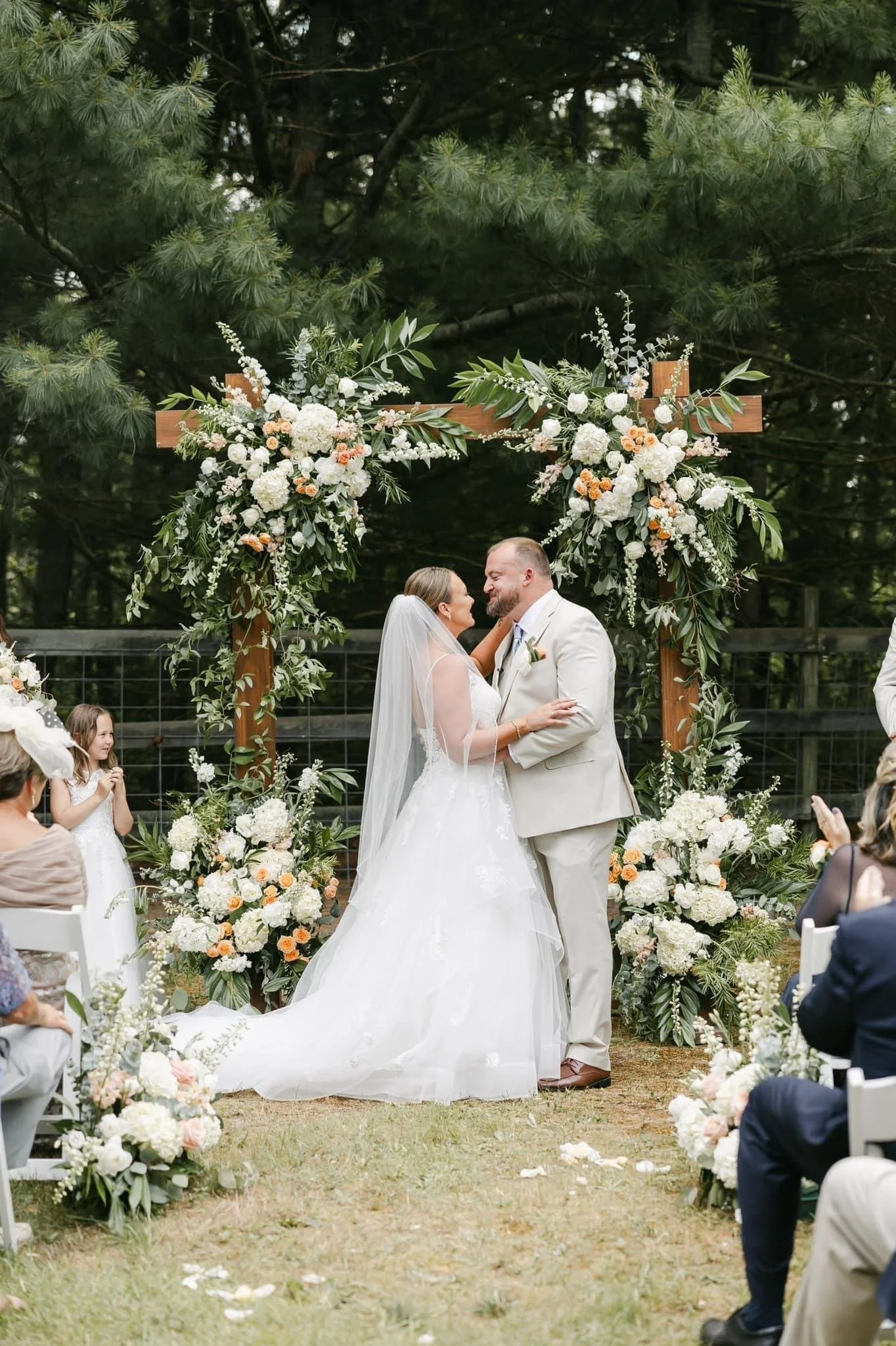 A bride and groom are sharing a kiss in an outdoor wedding ceremony. They are standing under a wooden arch adorned with white and peach flowers and greenery. People are seated around them, observing the ceremony, and a young girl in a white dress is 