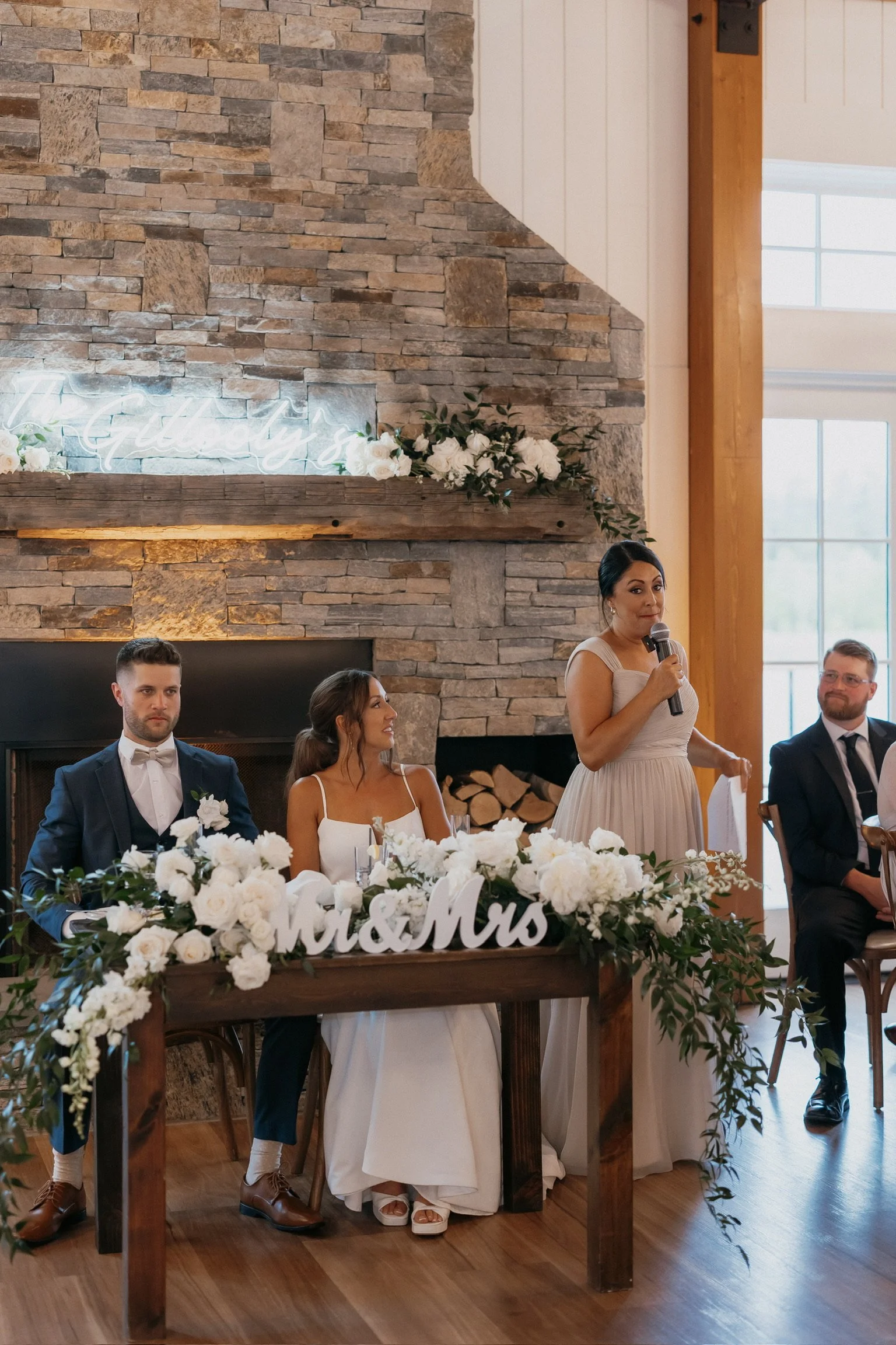 Wedding reception scene with a bride and groom sitting at a decorated table labeled "Mr & Mrs," while a woman stands giving a speech with a microphone, set against a stone fireplace backdrop.