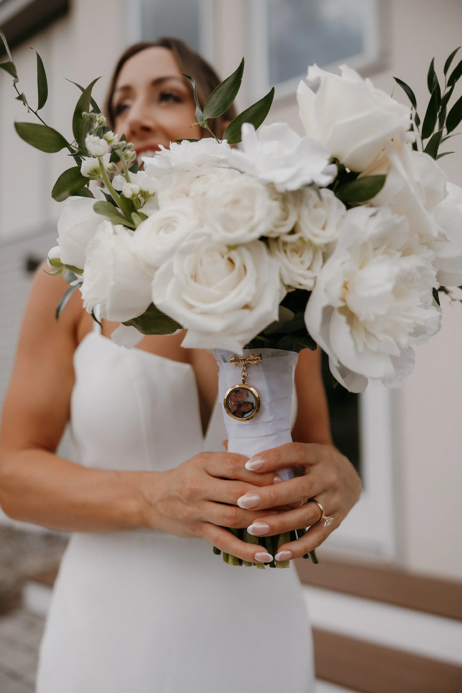 Bride holding a bouquet of white roses and greenery, with a circular locket attached to the bouquet's wrapping.