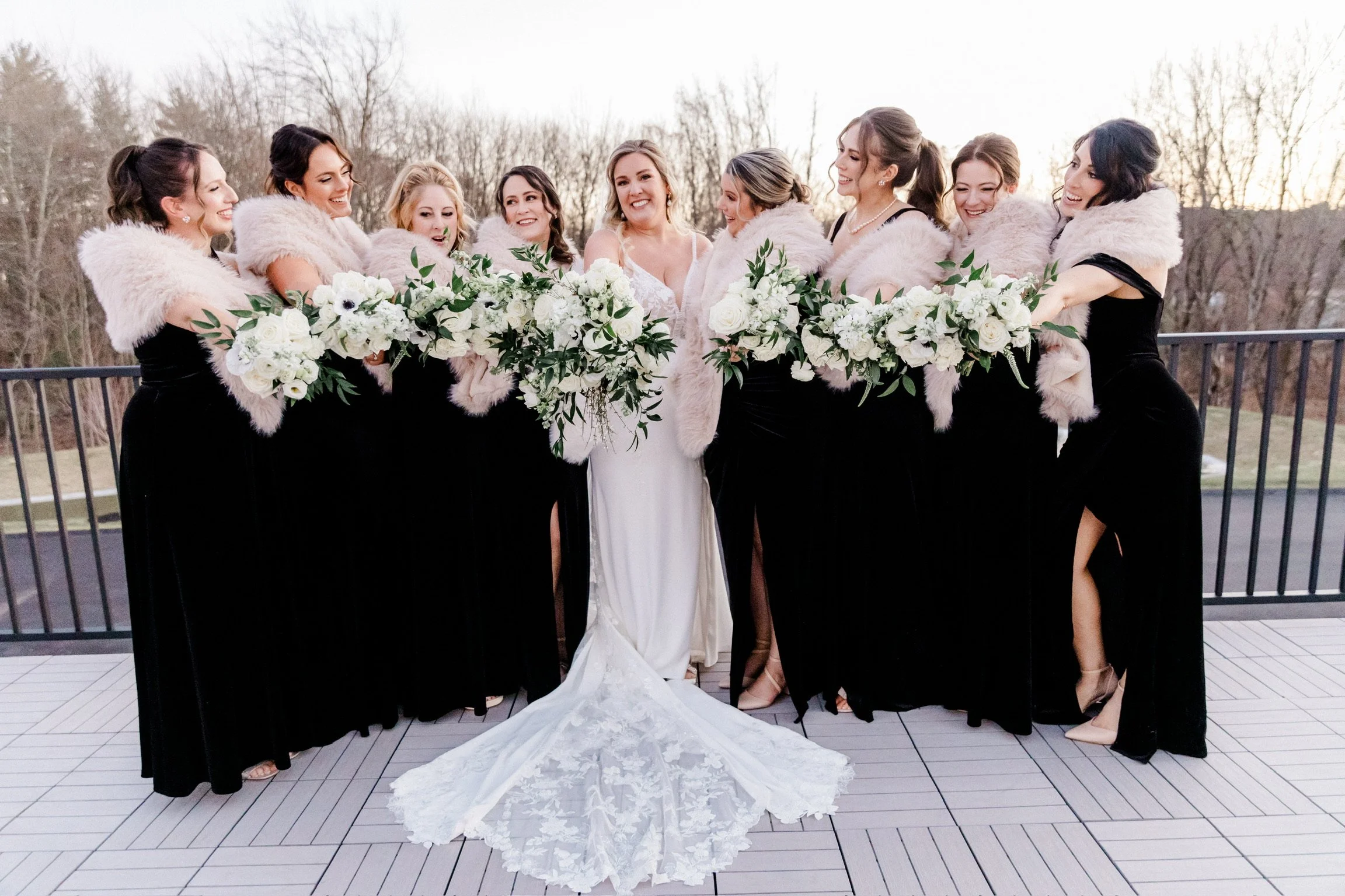 Bride and bridesmaids in black dresses and fur wraps holding white bouquets on an outdoor terrace.