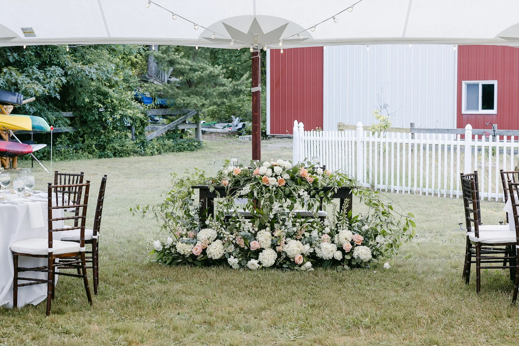Outdoor wedding reception setup with a floral arrangement on a table, under a tent canopy with string lights; white fence and red barn in the background.