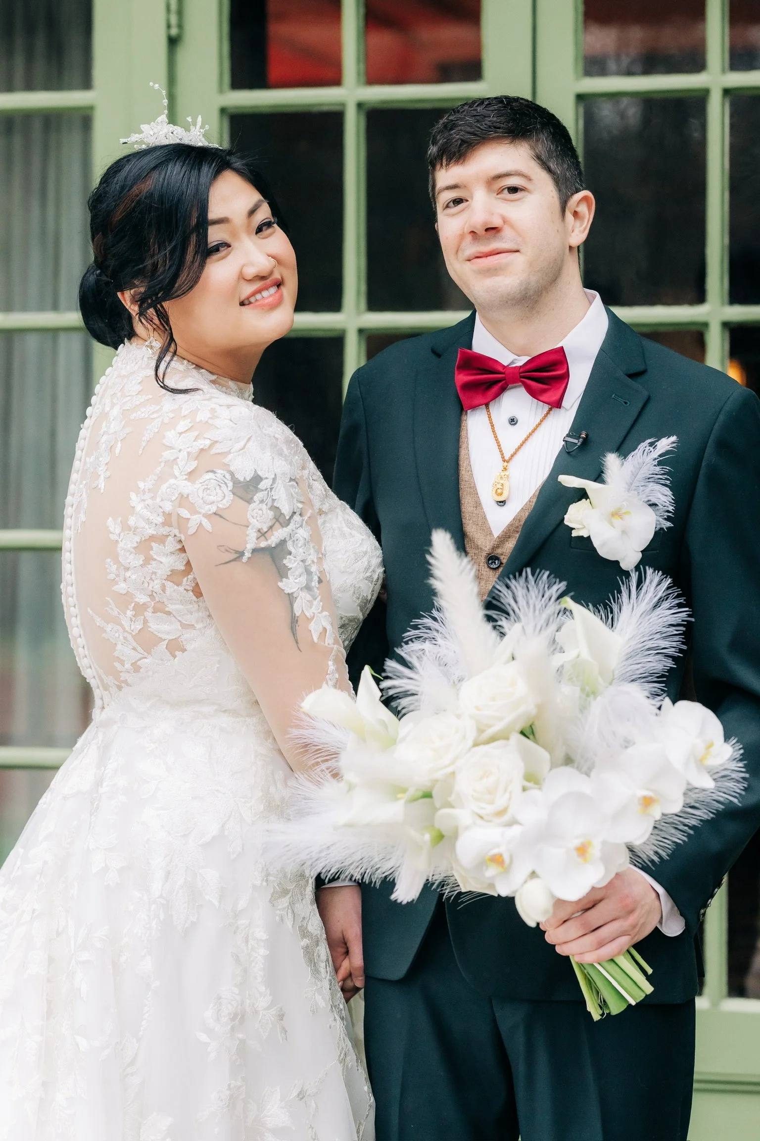 A bride and groom pose for a wedding photo. The bride wears a lace wedding dress with floral details, while the groom is in a suit with a red bow tie. They are holding a bouquet of white flowers. In the background, there is a green window frame.