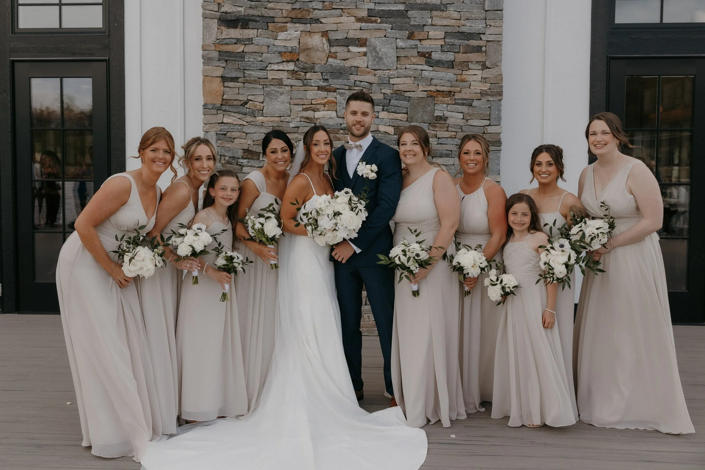 Wedding party photo with bride, groom, bridesmaids, and flower girls in light dresses and dark suit, holding white bouquets, standing in front of stone wall.