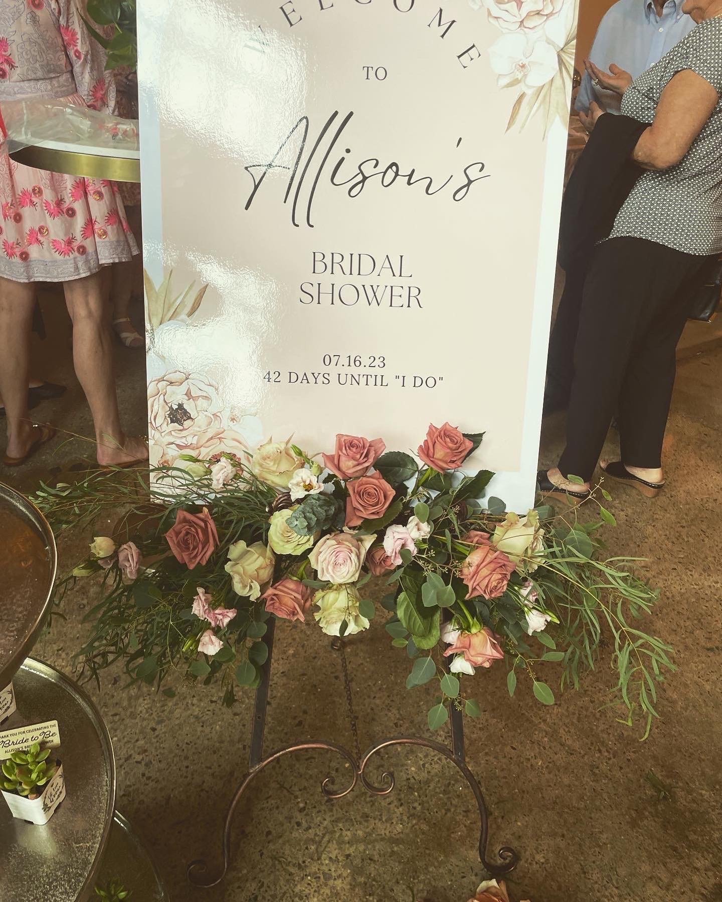 Bridal shower welcome sign with floral arrangement, reading "Welcome to Allison's Bridal Shower, 07.16.23, 42 days until 'I do'," surrounded by people.