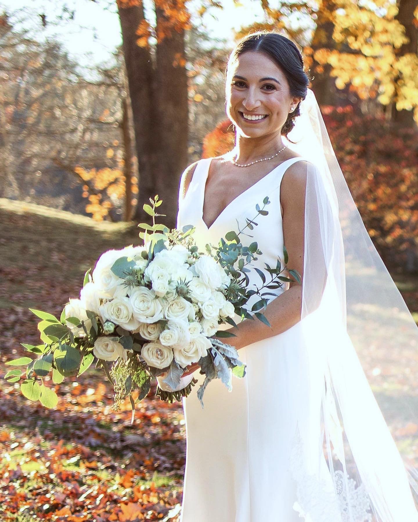 Bride in white dress holding a bouquet of white roses and greenery, standing outdoors with autumn foliage in the background.