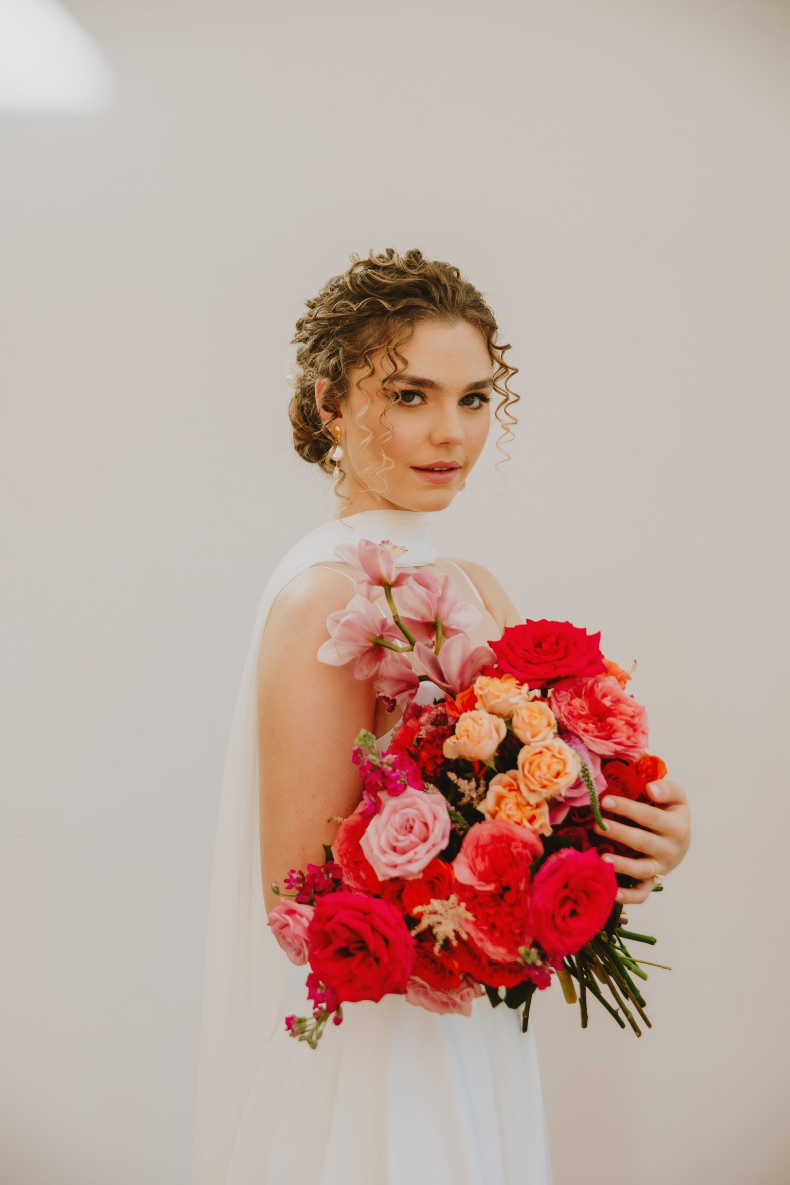 Woman in white dress holding colorful bouquet of roses and lilies against a plain background.