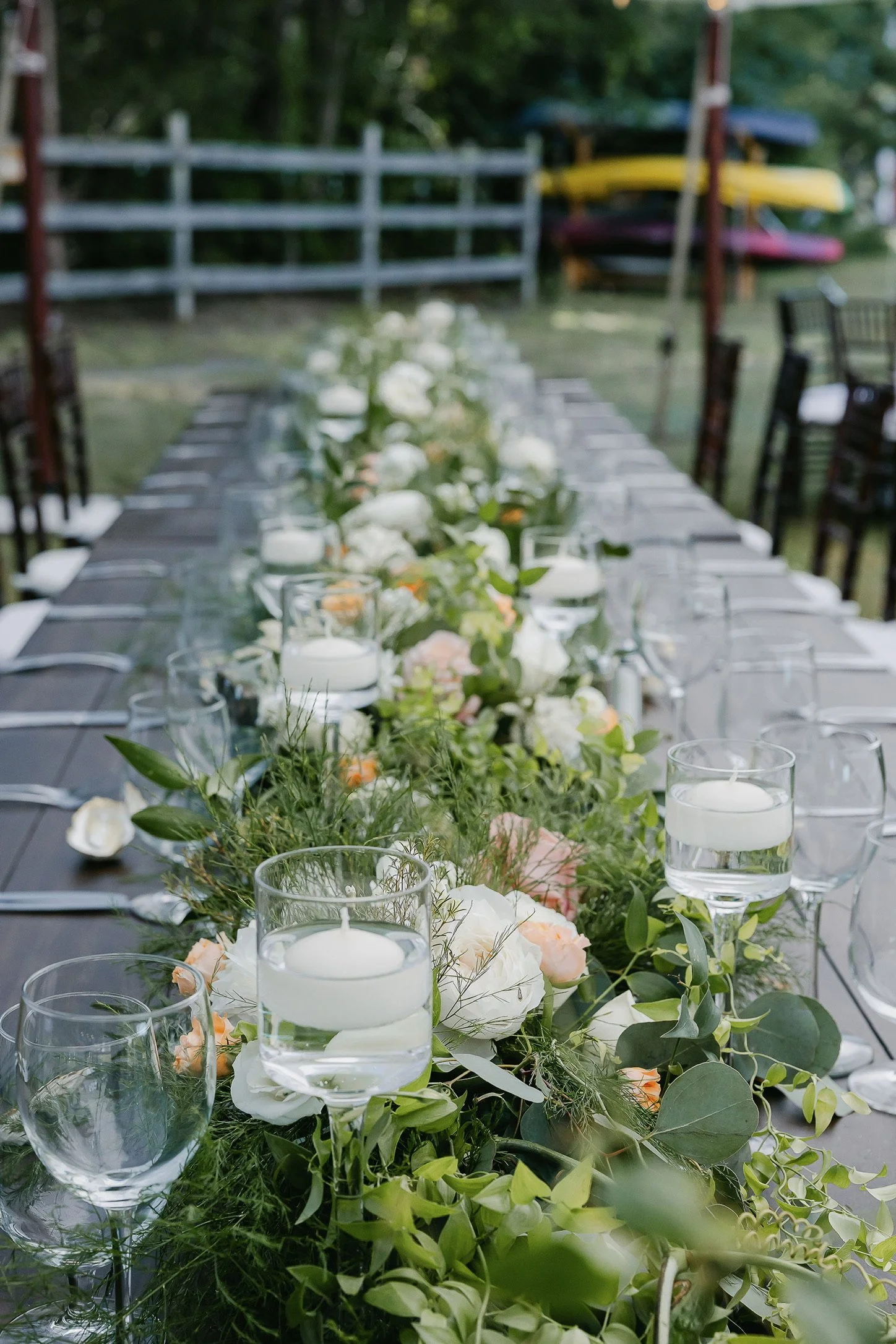 Long outdoor dining table with floral centerpiece, greenery, white and peach flowers, float candles in glass holders, and wine glasses set for an event.