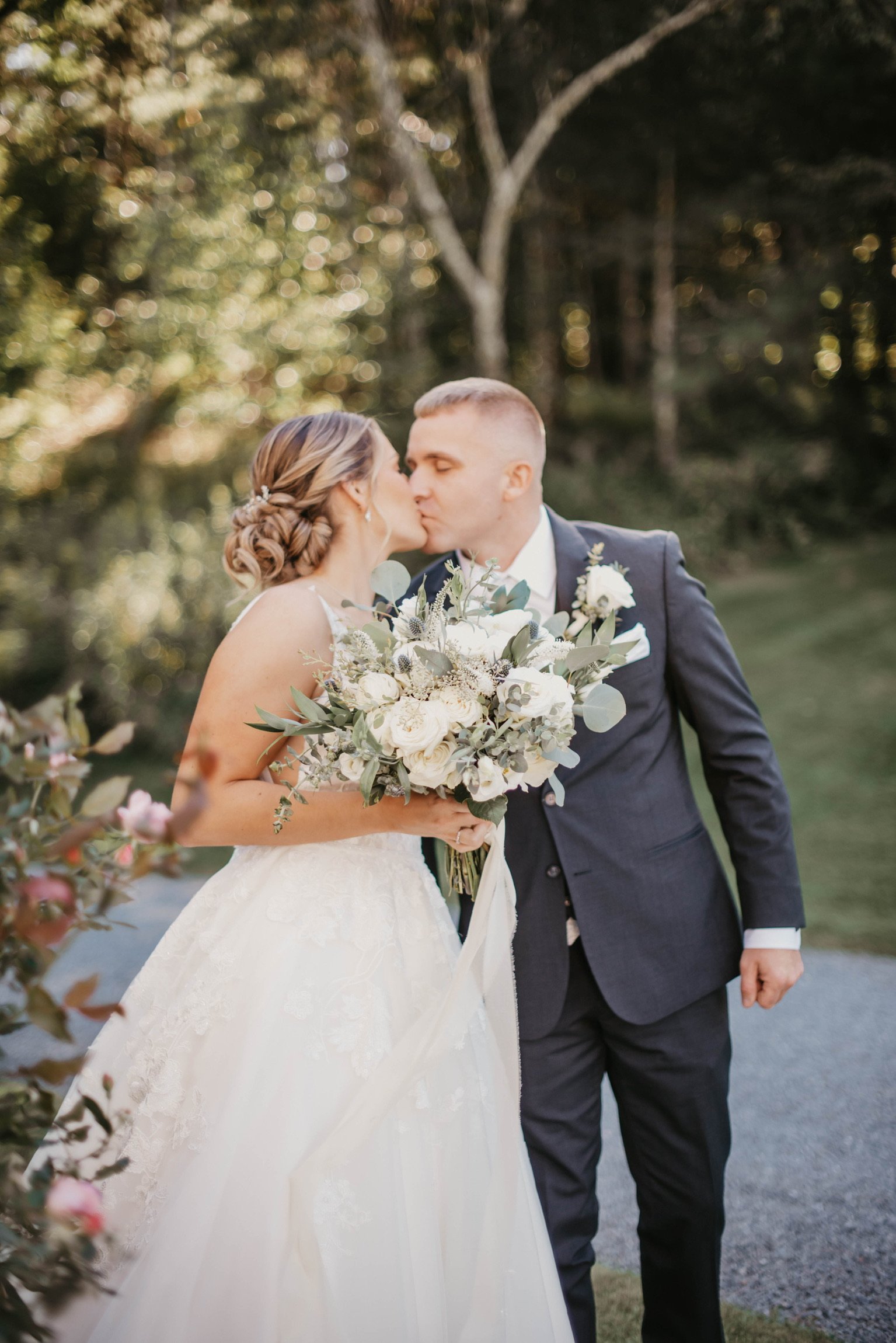 A bride and groom share a kiss outdoors, with the bride holding a bouquet of white flowers and greenery, and the groom in a dark suit.