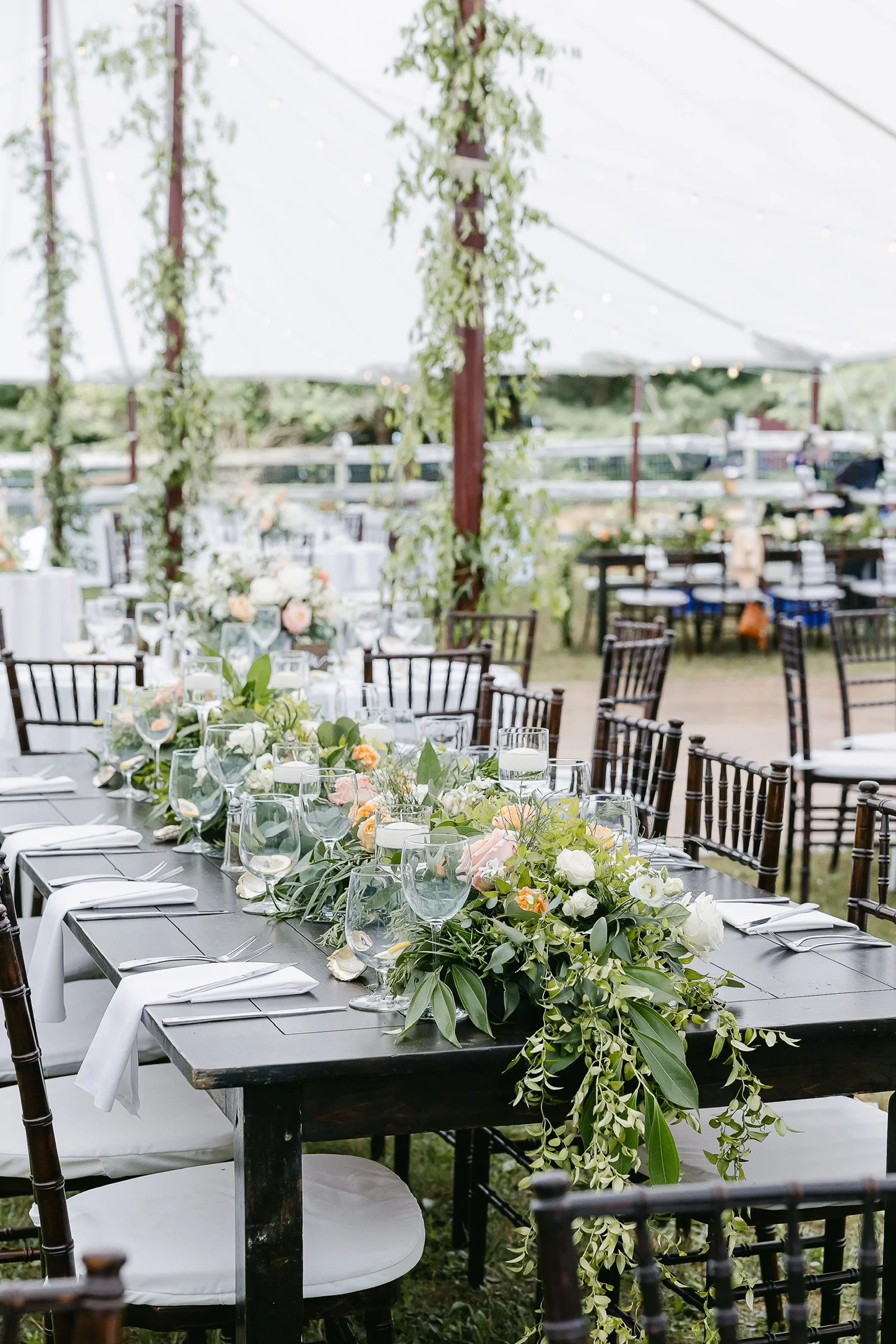 Elegant outdoor table setting under a tent with floral centerpiece, green foliage, and wine glasses on a wooden table surrounded by chairs.