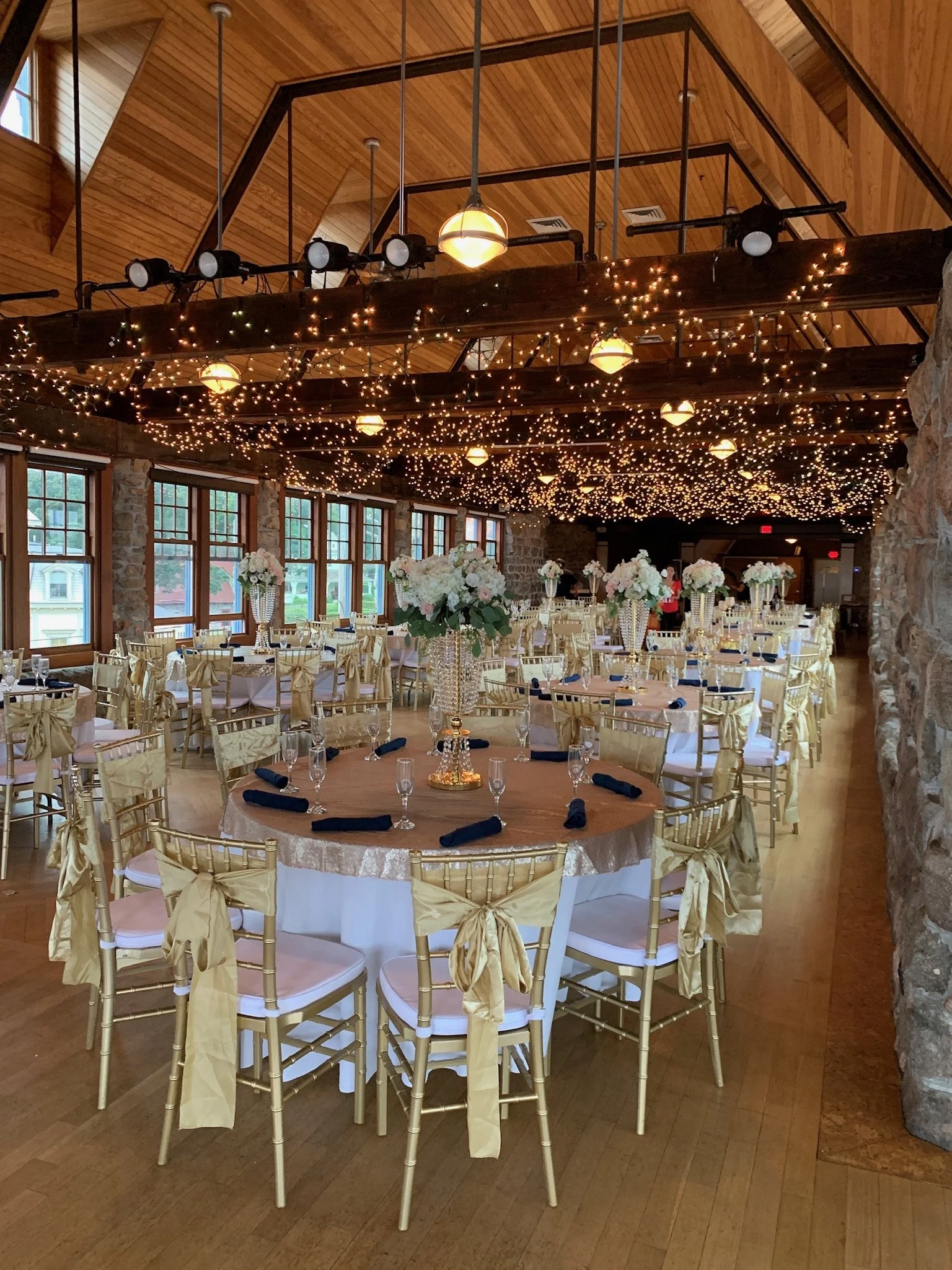 Elegant reception hall with round tables, gold chairs, floral centerpieces, and string lights on wooden beams in a rustic venue.