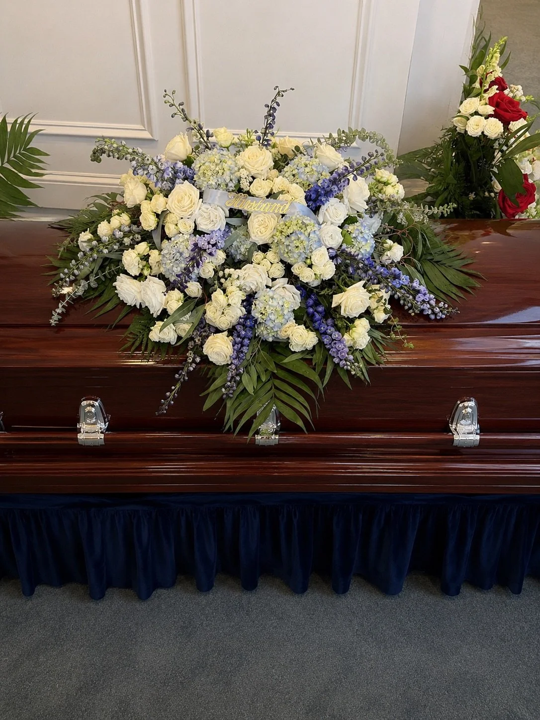 A closed wooden casket with a large floral arrangement on top, featuring white roses, purple delphiniums, and green foliage. A small sign with the name 'Auburn' is in the center of the flowers. The casket rests on a dark blue cloth beneath a white wa