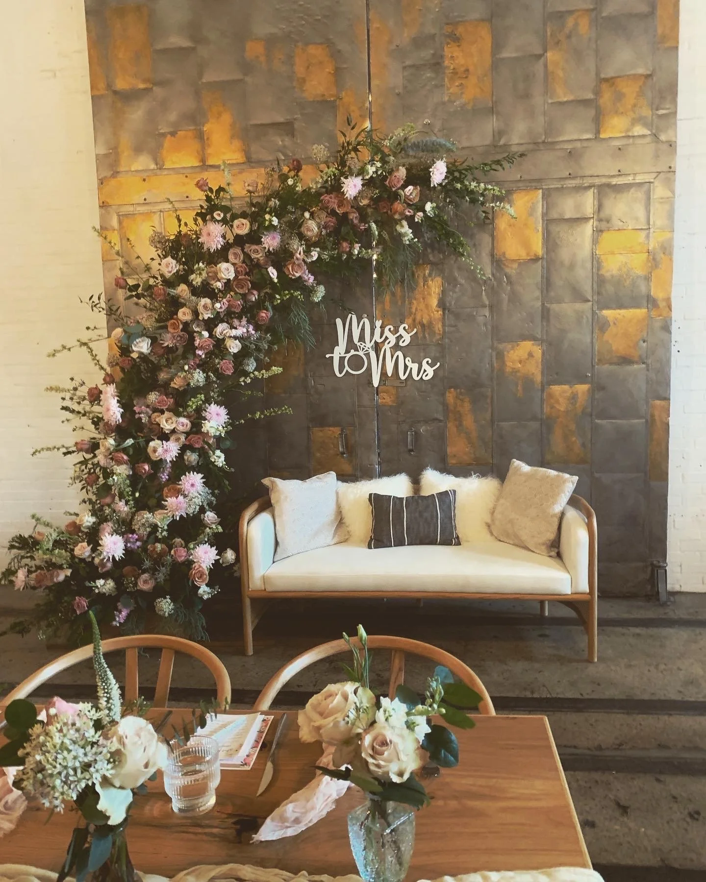 Wedding-themed decor with a white couch, floral arrangement, and "Miss to Mrs" sign, in front of a decorative wall, with a wooden table set with flowers in vases.