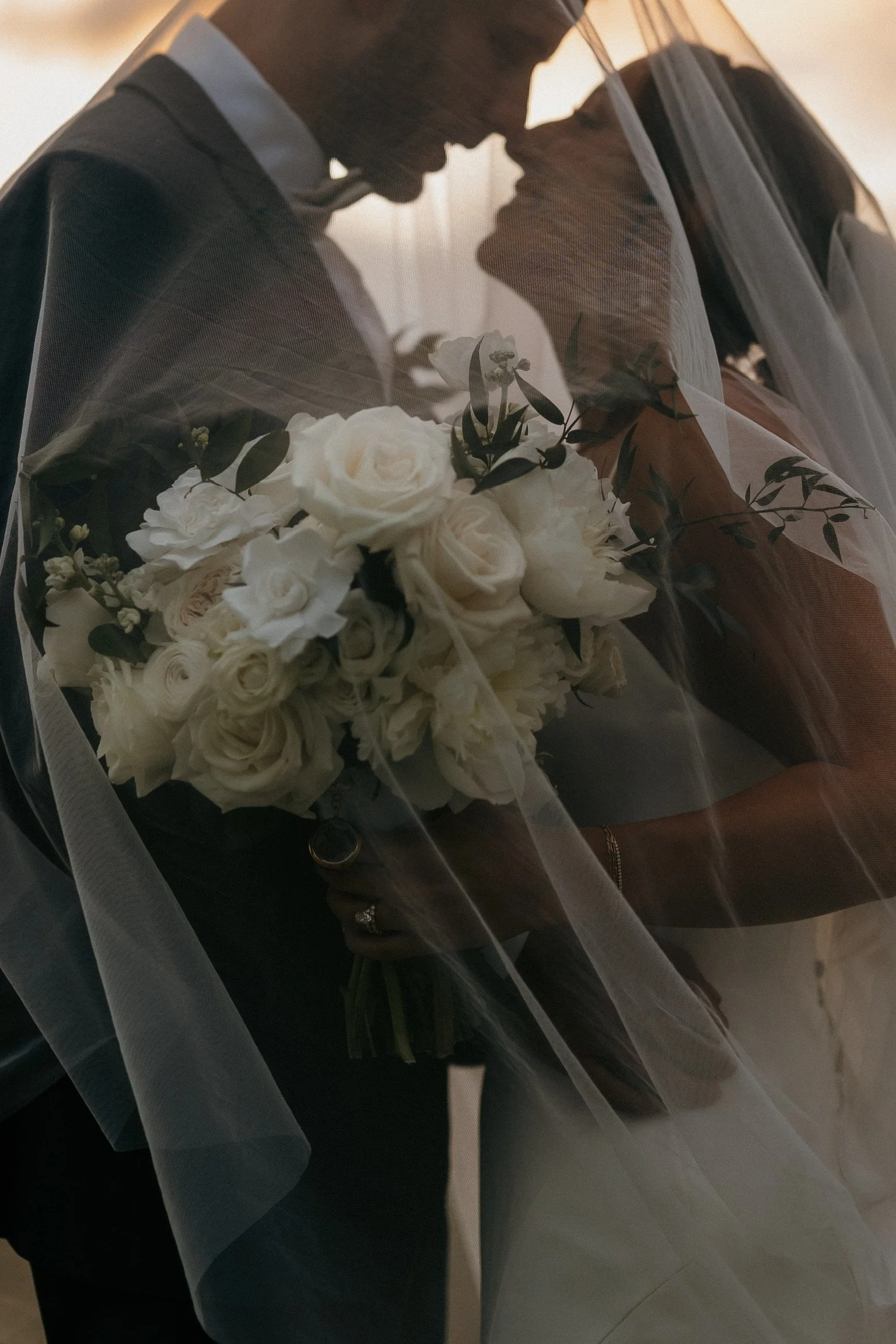 Bride and groom embracing under a veil with a bouquet of white roses.
