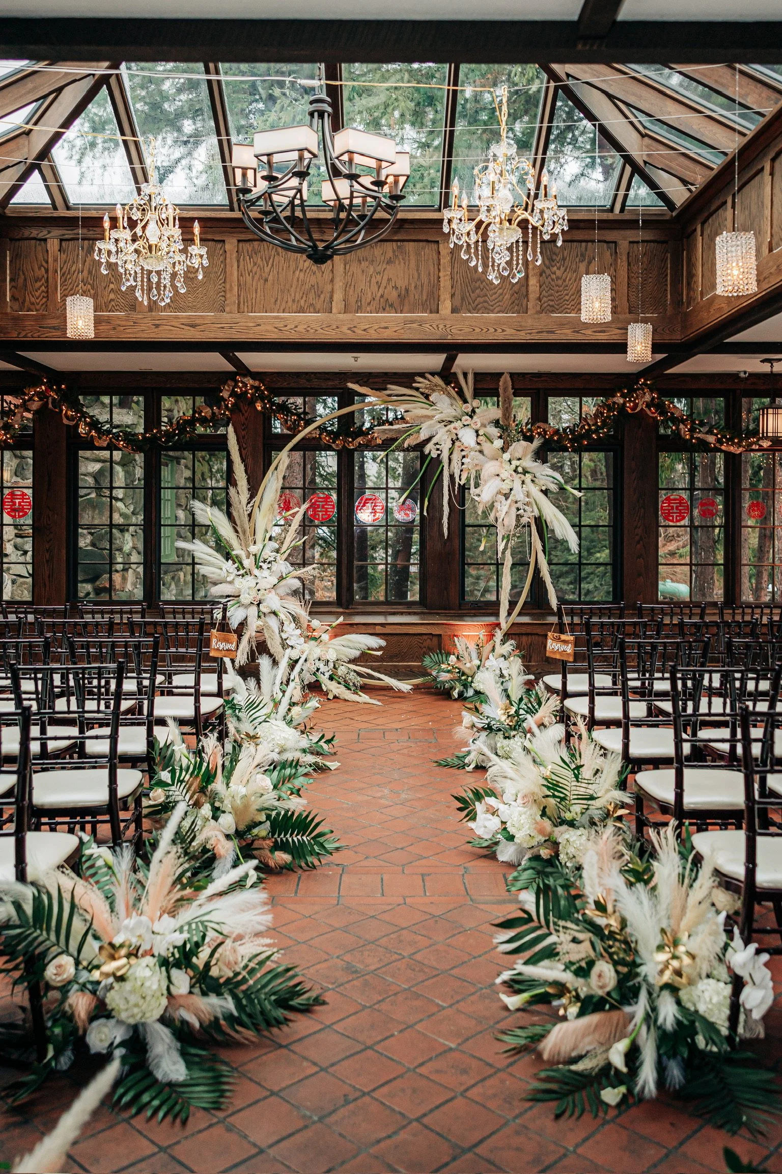Indoor wedding ceremony setup with floral decorations, elegant chandeliers, and arranged chairs. The aisle is adorned with greenery and white flowers, leading to a floral arch under a glass ceiling.
