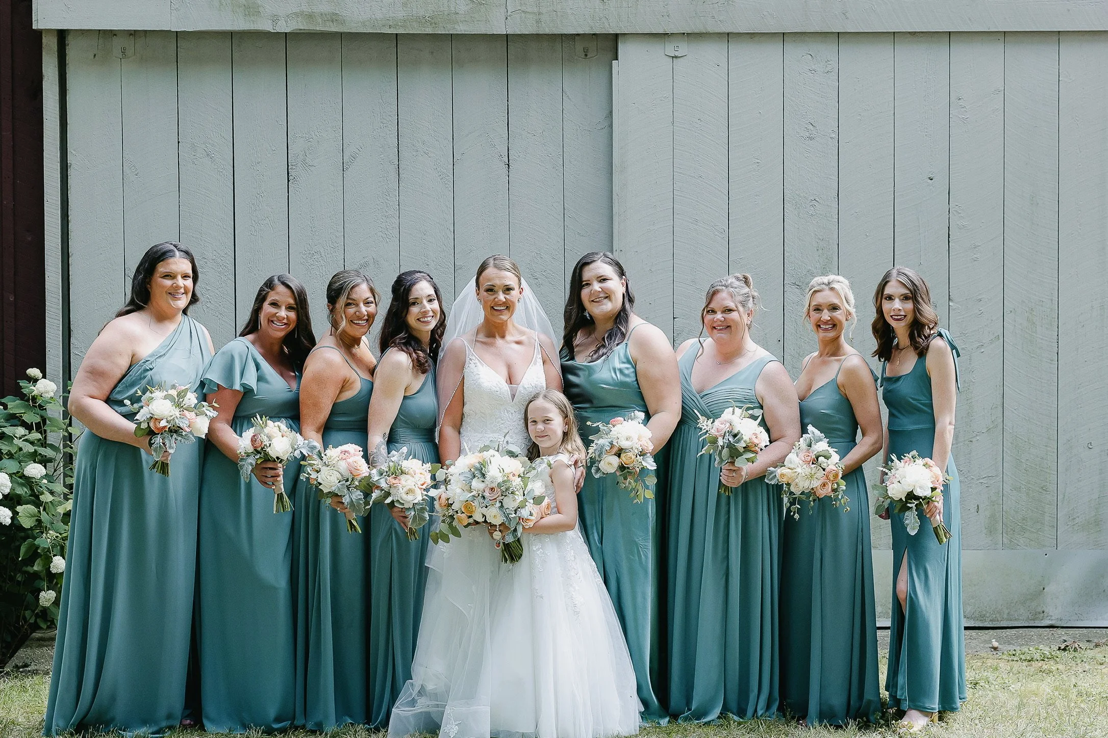A bride and her bridesmaids in teal dresses standing outdoors in front of a wooden wall, all holding bouquets of flowers.